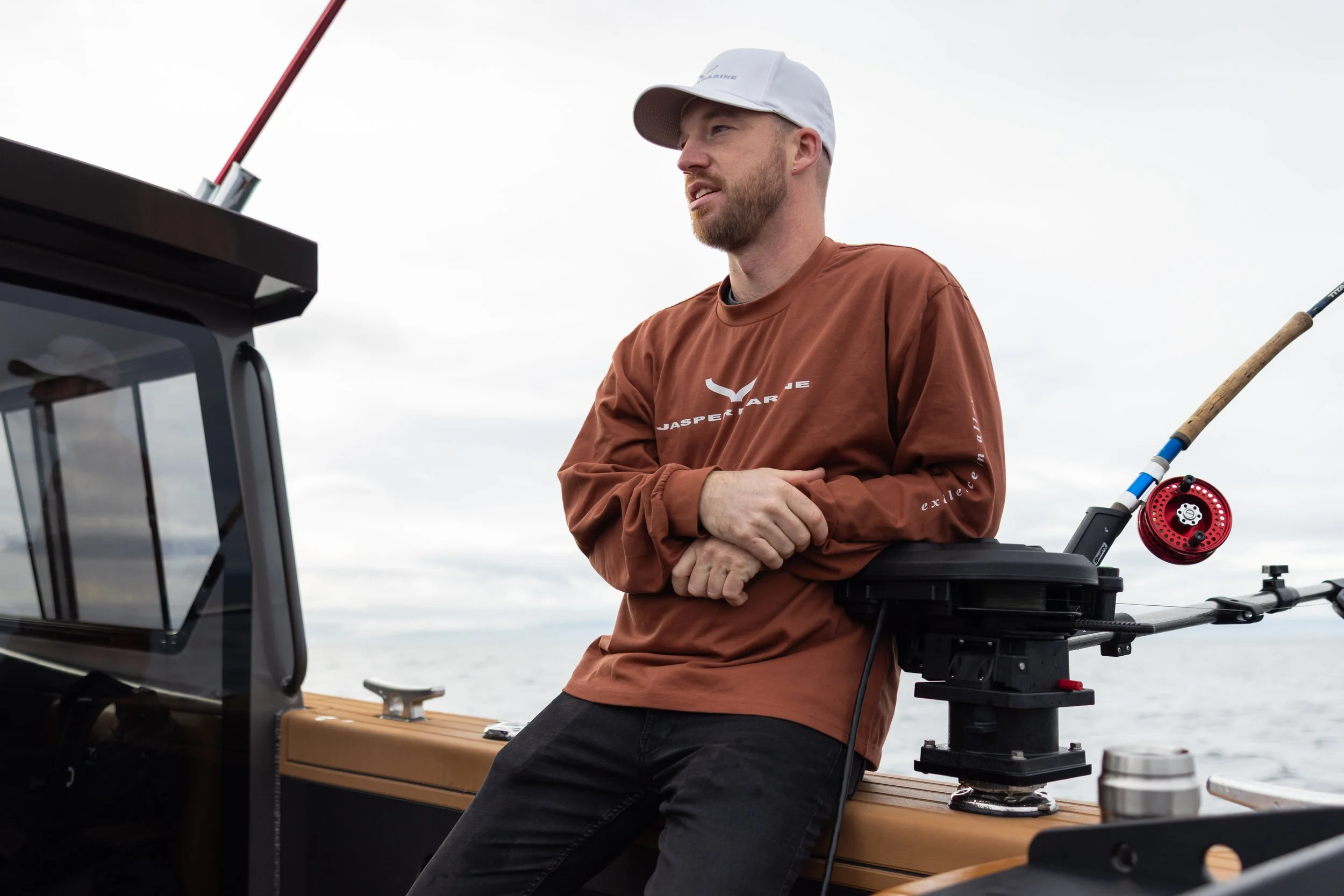 Jasper of Jasper Marine wearing a white cap and a brown sweater, leaning on a boat railing with fishing gear, looking out at the water with a cloudy sky in the background.