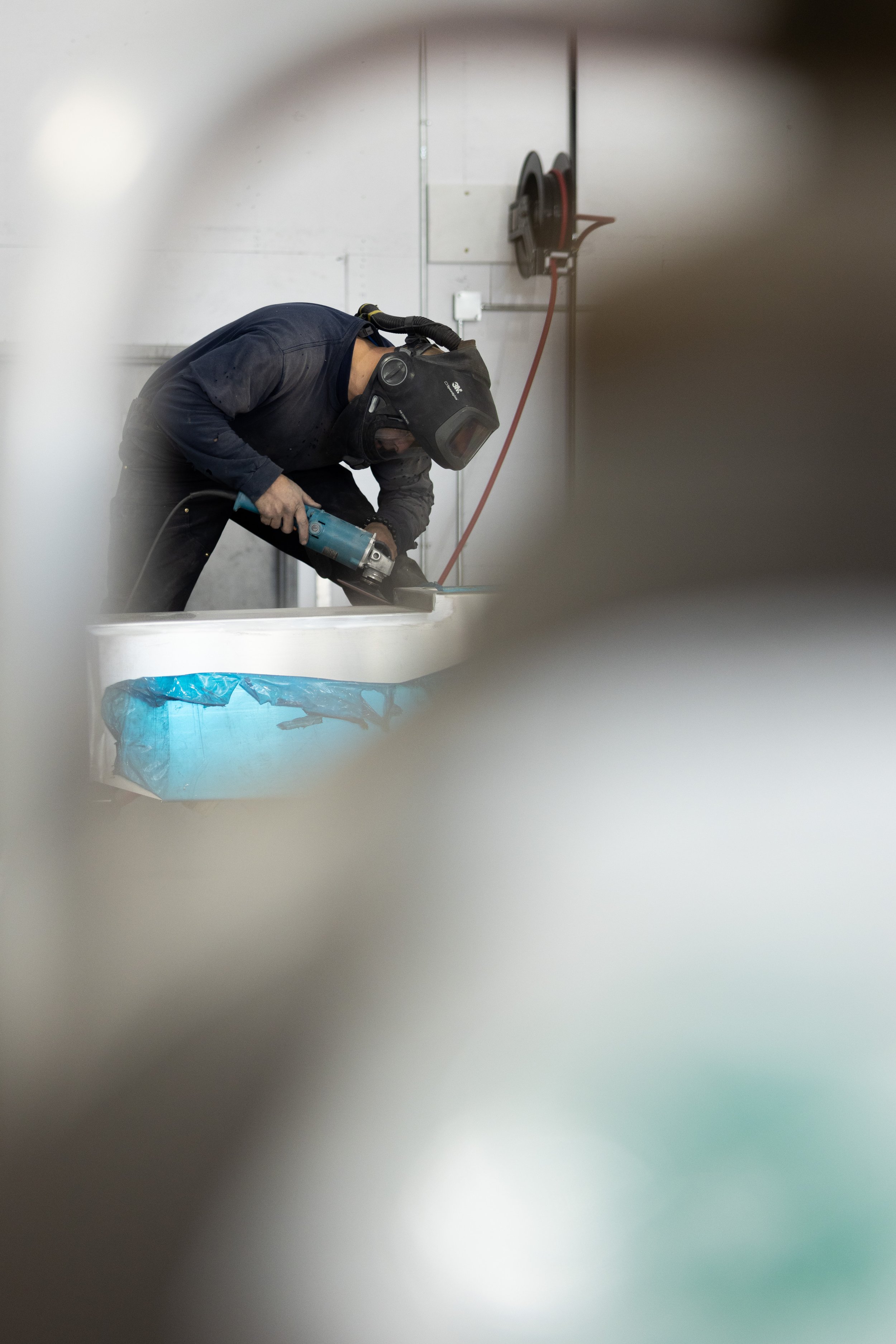 A welder wearing a mask and protective gloves, using a tool on a surface in an industrial setting, with a hose reel on the wall in the background.