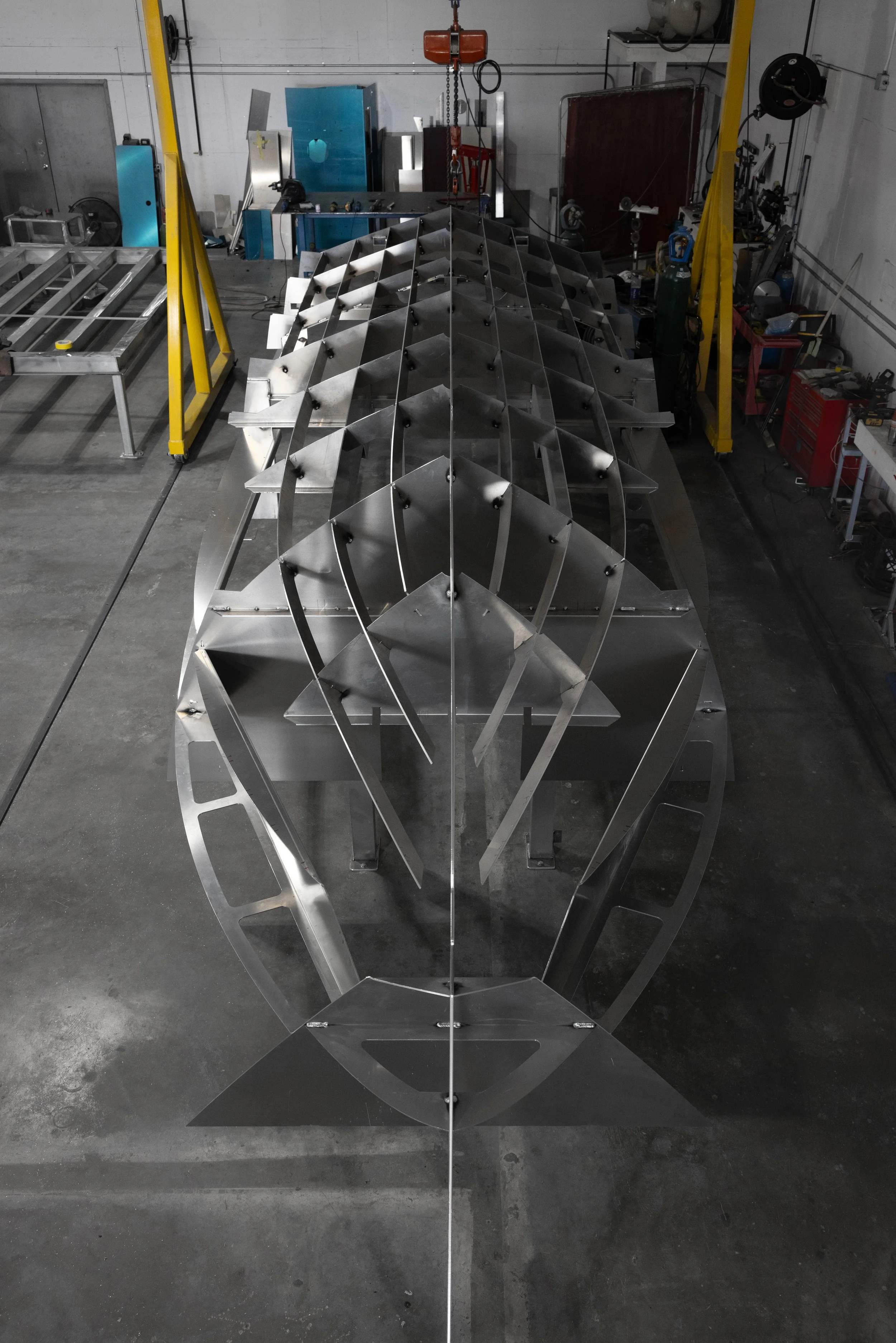 Aluminum boat hull under construction in a workshop with machinery and tools in the background.