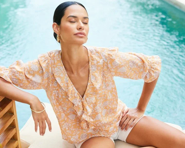 A woman relaxing by a pool, wearing a peach-colored floral blouse and white shorts, with eyes closed and a serene expression.