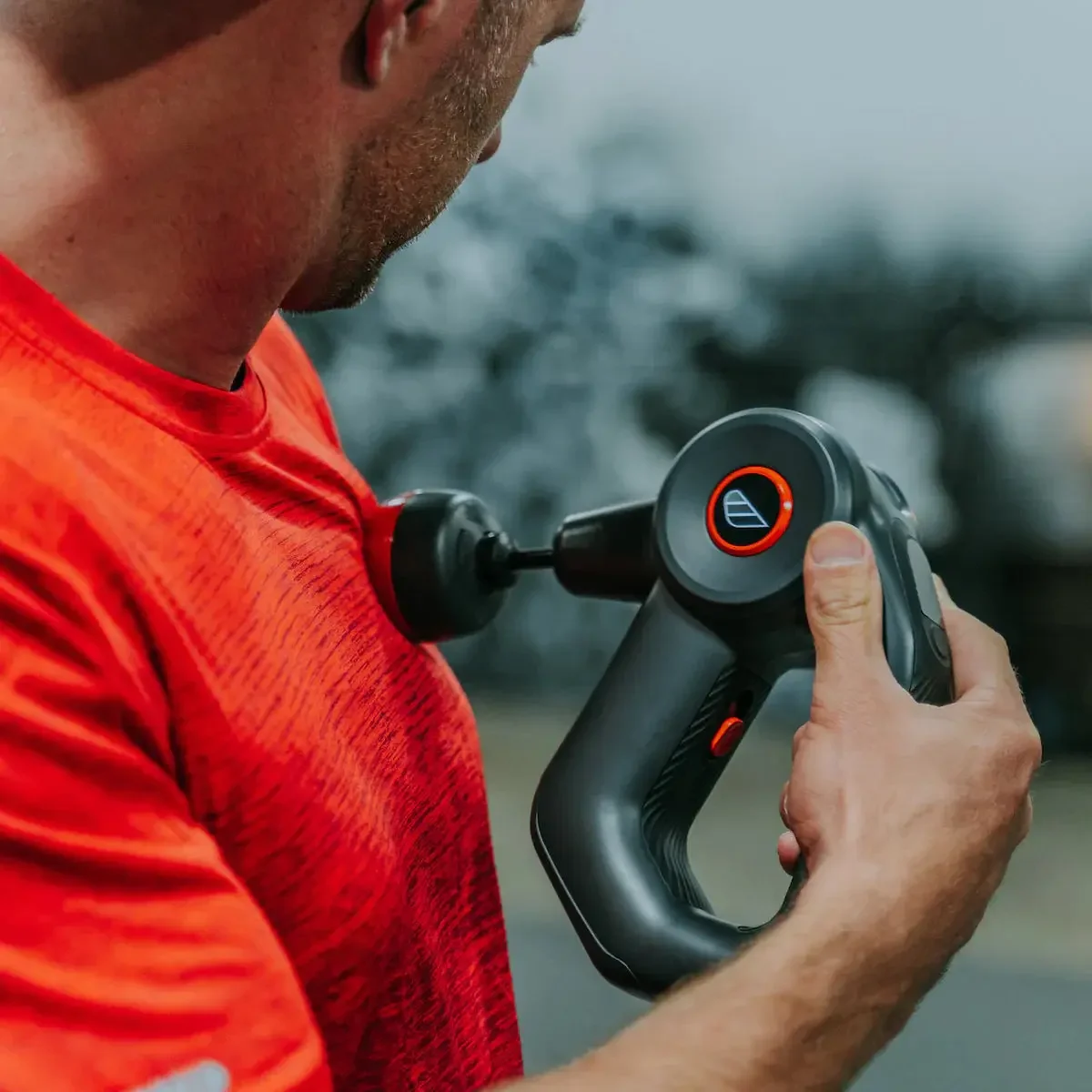Close-up of a man in a red shirt using a massage gun on his shoulder, outdoors with blurred background.
