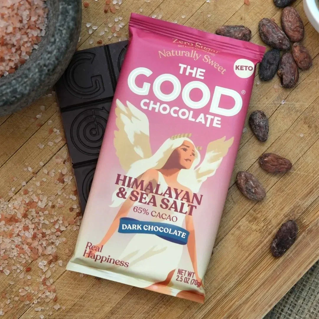 A pink package of The Good Chocolate with Himalayan and sea salt, dark chocolate flavor, 65% cacao, on a wooden surface surrounded by cacao beans and Himalayan salt crystals.