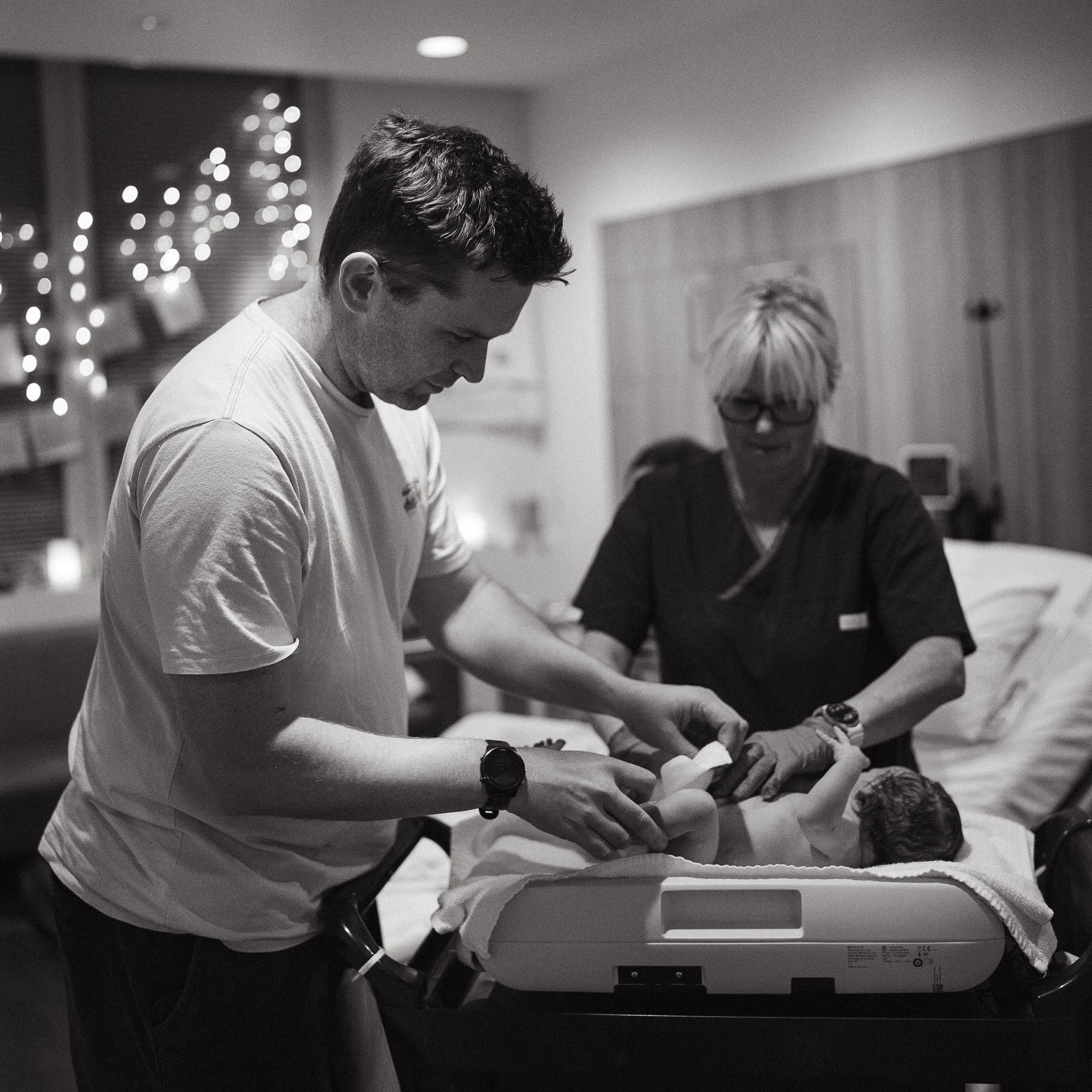 A young man with curly hair and a wristwatch, wearing a light-colored t-shirt, is leaning over a hospital bed, interacting with a woman lying in the bed. An elderly woman with short light-colored hair and glasses is also present, observing the interaction.