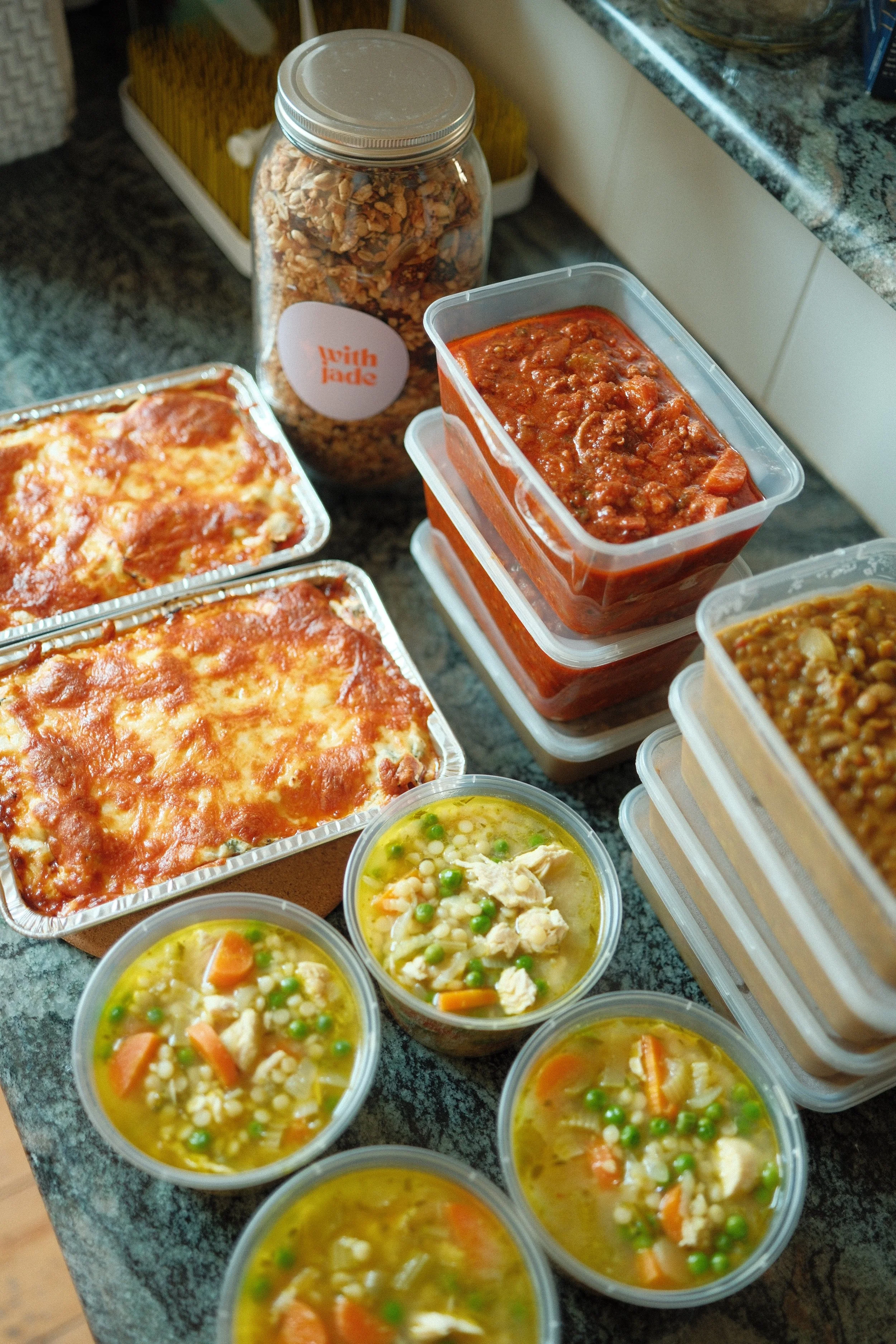 Several containers of prepared food, including lasagna, chili, lentil stew, and chicken noodle soup, are arranged on a kitchen countertop next to a jar of granola.