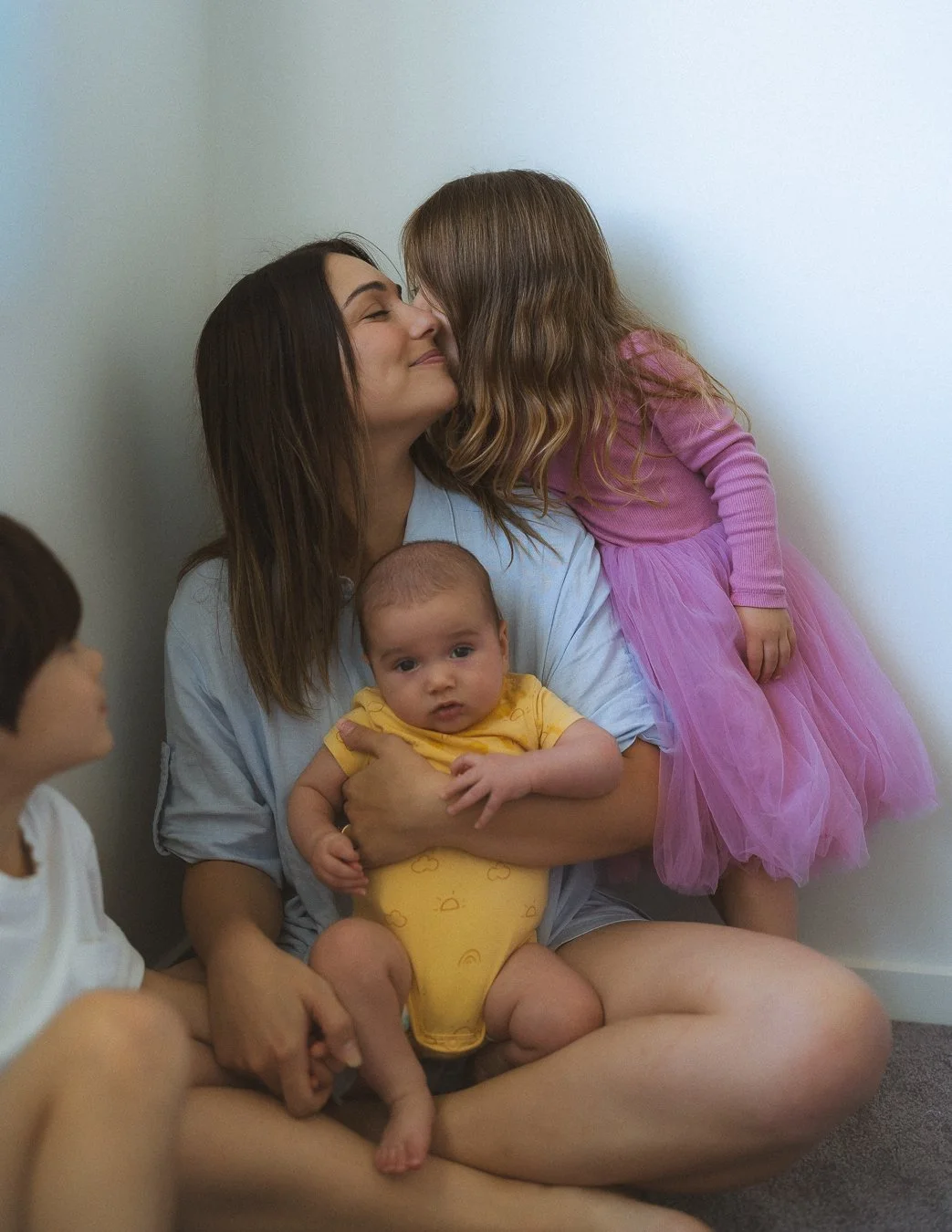 A woman is sitting cross-legged with three children in a corner, one of whom is leaning in to give her a kiss. The woman is holding a baby wearing a yellow outfit, and another child wearing a pink dress is standing on her lap, leaning in for a kiss. A third child is sitting nearby, out of focus.