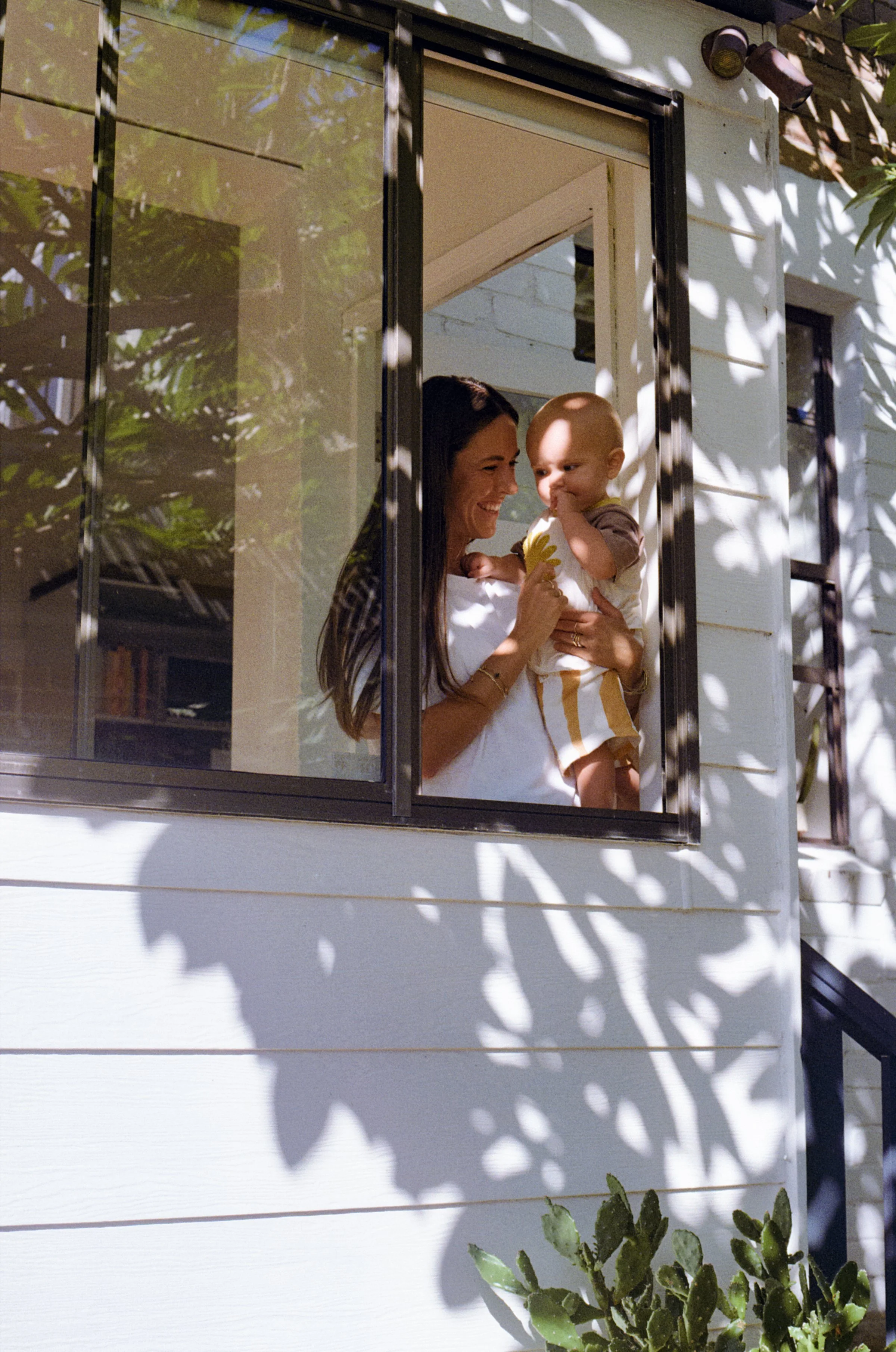 A woman and a baby are smiling and looking at each other through a window. The woman holds the baby in her arms, and they are both smiling happily. The window has shadows of leaves cast on it. There are some green plants below the window.