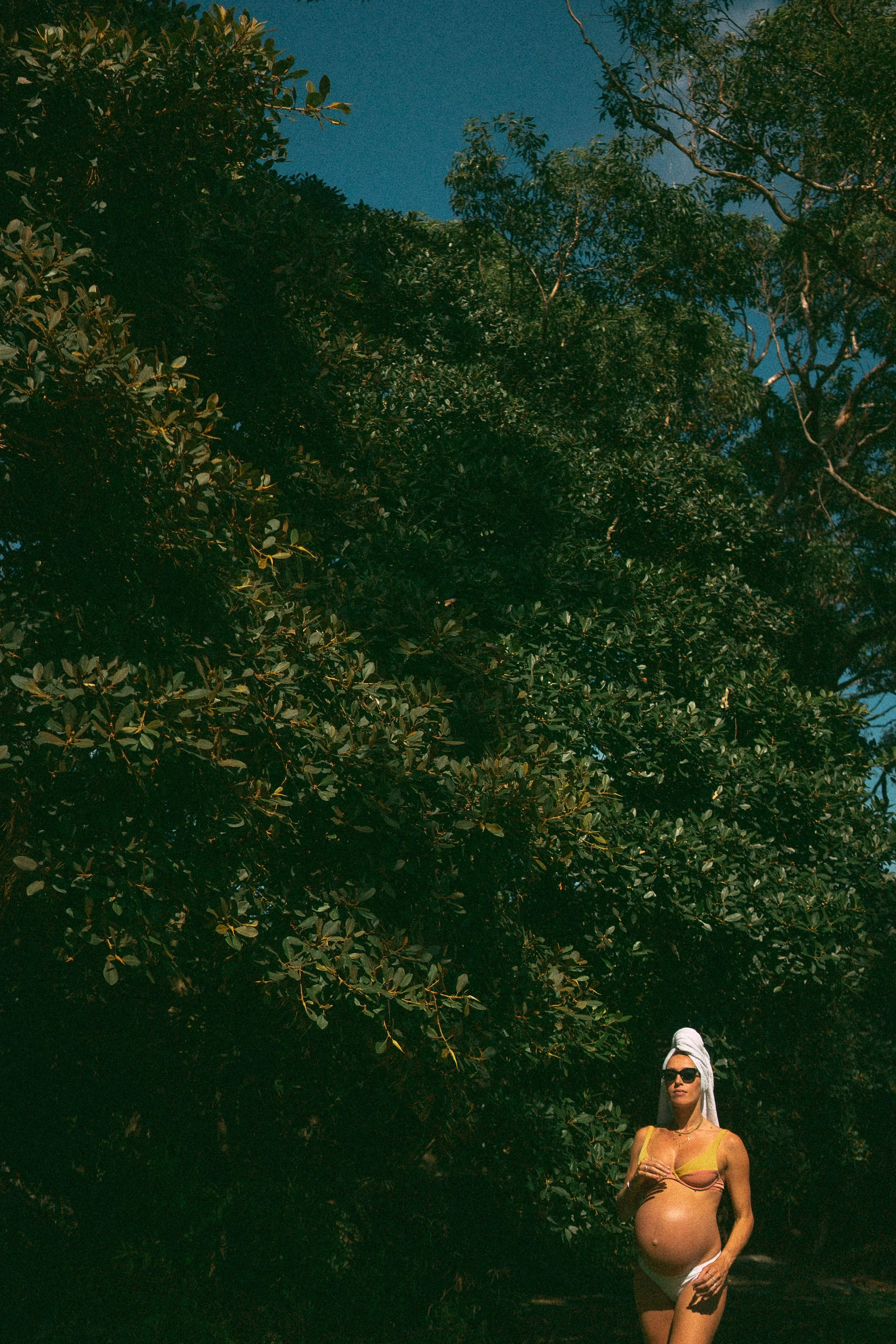 A woman with sunglasses wearing a yellow top and white shorts, pregnant, standing outdoors beneath large leafy green trees at night.