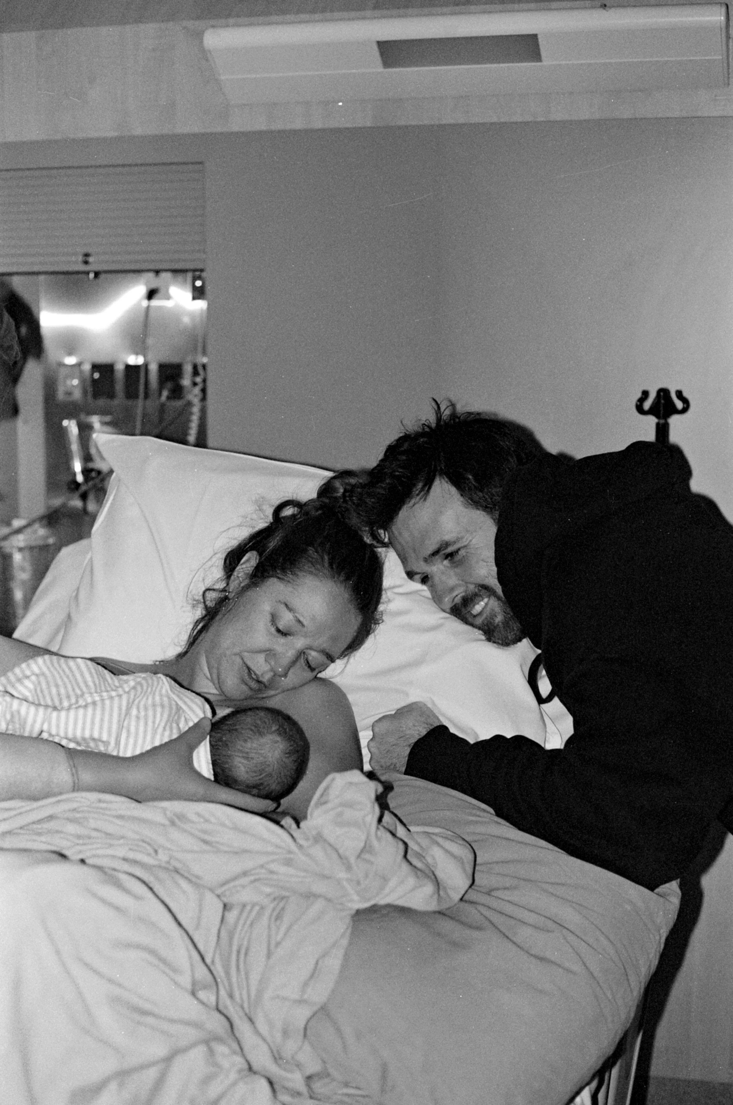 A woman lying in a hospital bed breastfeeding a newborn, with a man beside her leaning over and smiling, in a hospital room.