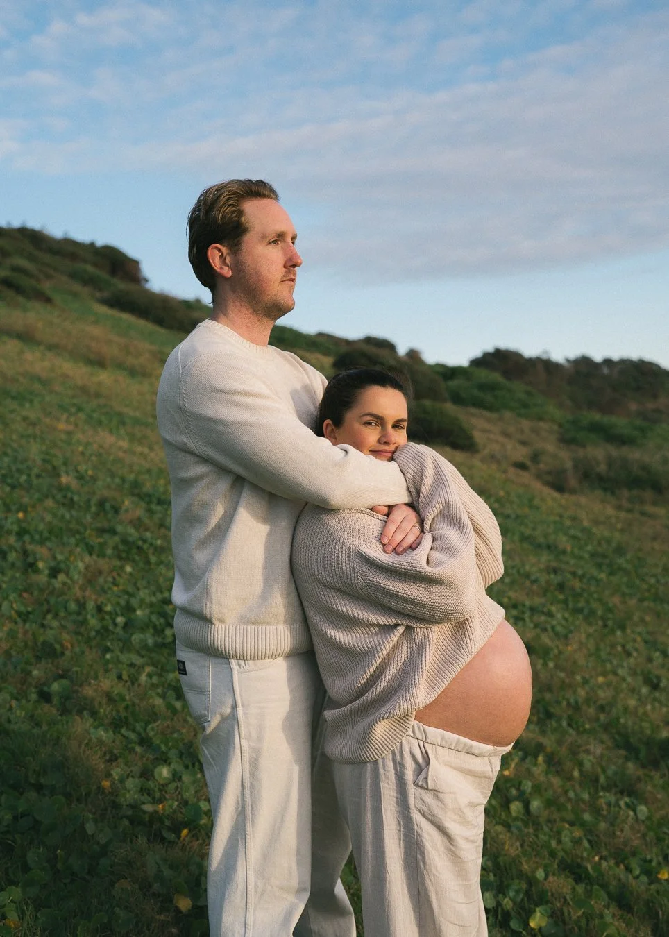 A man and a pregnant woman standing outdoors on a grassy hillside, with the woman hugging her belly and smiling at the camera, while the man looks into the distance.