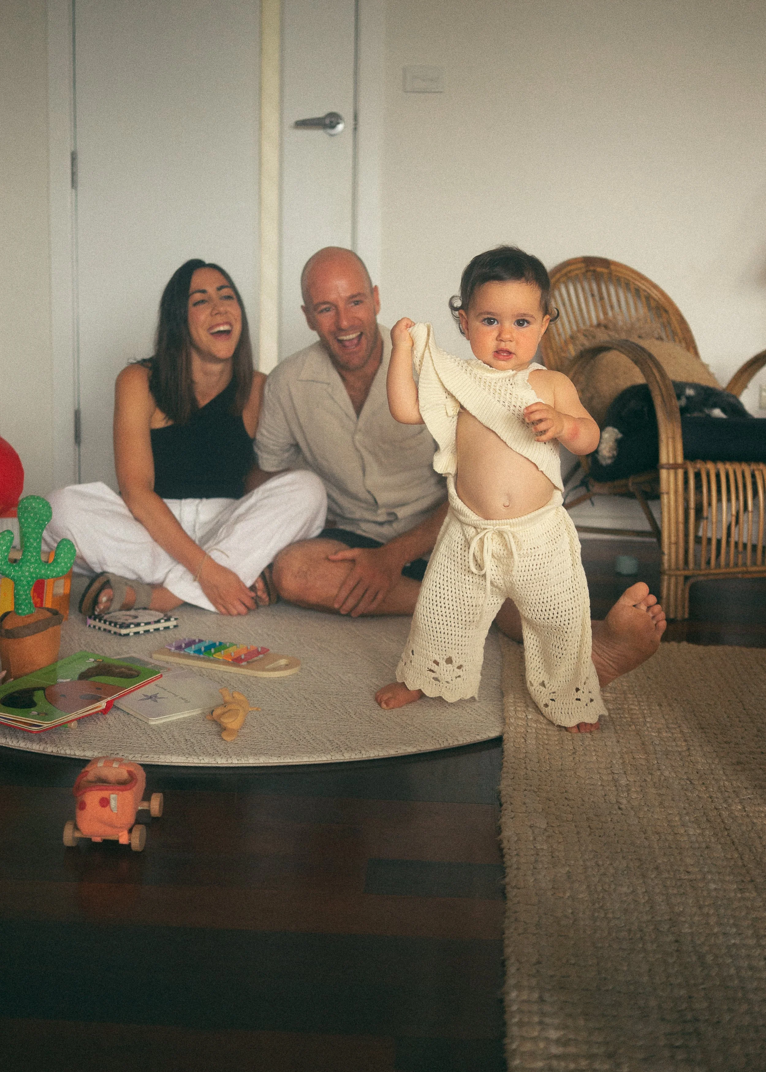 A young girl in cream-colored crochet pants and a top, standing on a rug, lifting her shirt to reveal her belly, with two adults, a man and a woman, sitting on the floor behind her, laughing and smiling. Toys and books are scattered around on the rug.
