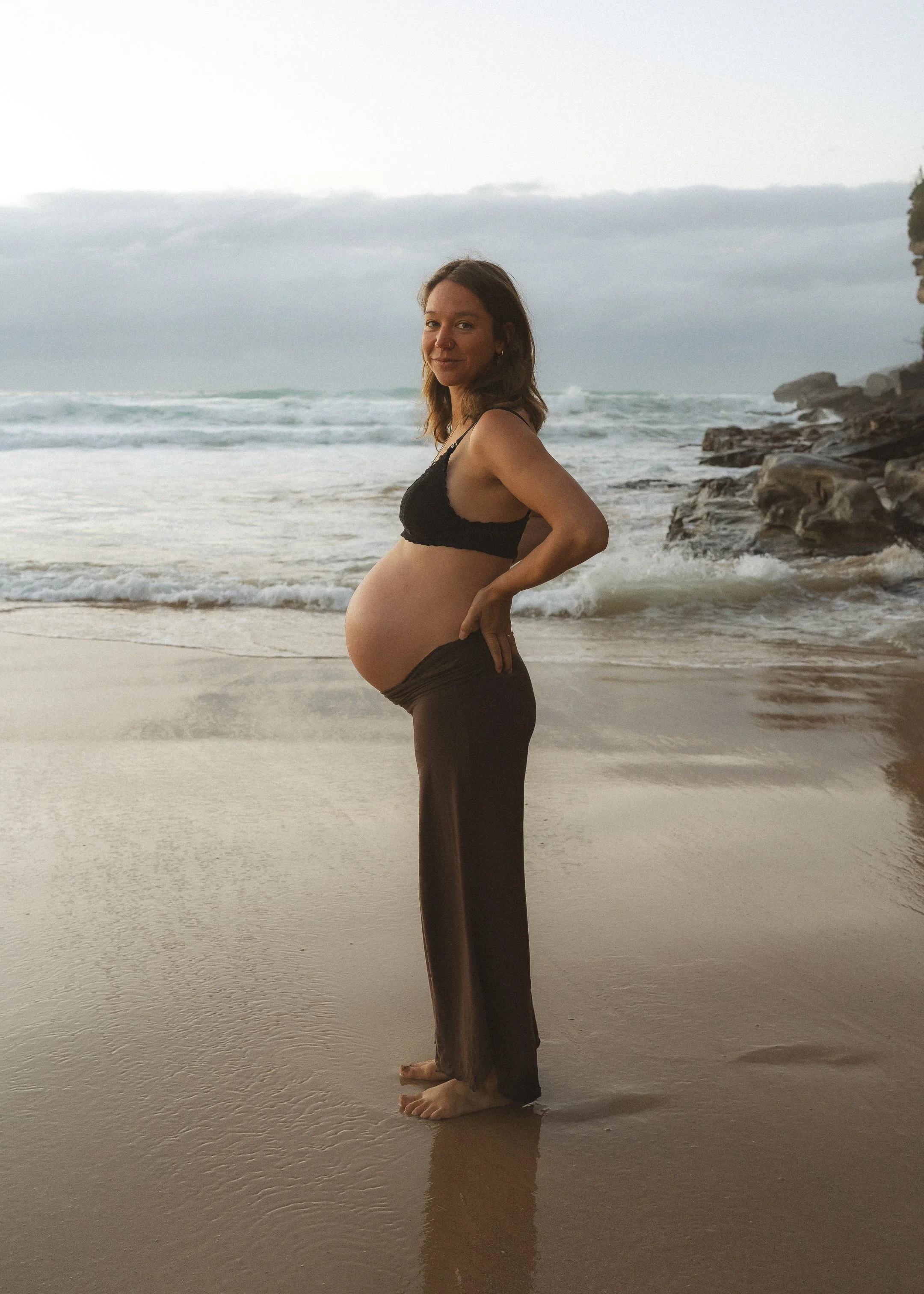 A woman standing on a beach. Her pregnant belly is proudly displayed, her feet are bare, the sky is pale blue and grey behind her