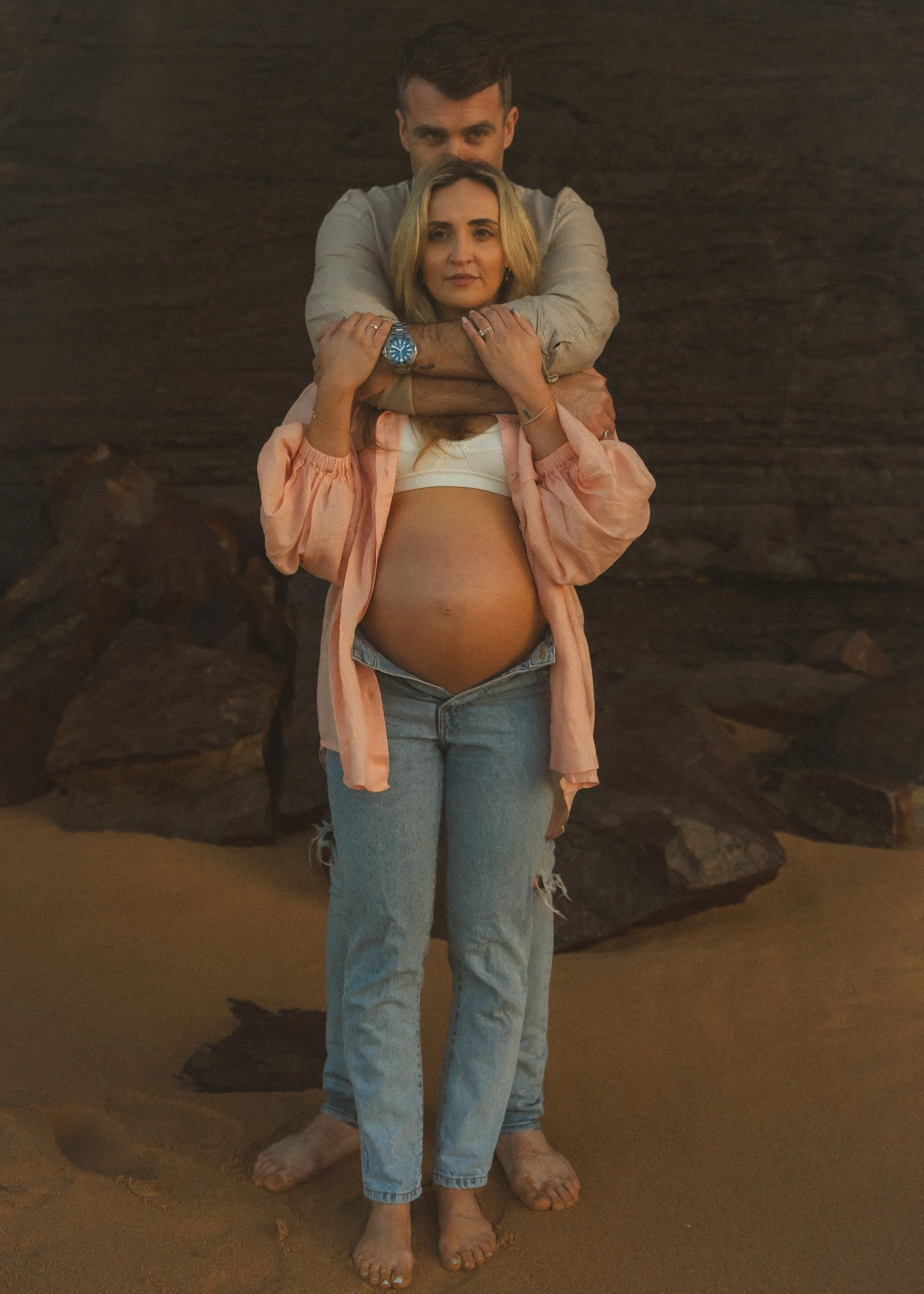 A couple standing on a sandy beach, with rocky formations in the background. The woman is pregnant and wearing a light pink jacket, a white crop top, and ripped jeans. The man stands behind her with his arms around her shoulders, both barefoot, and the woman’s feet are placed at their feet.