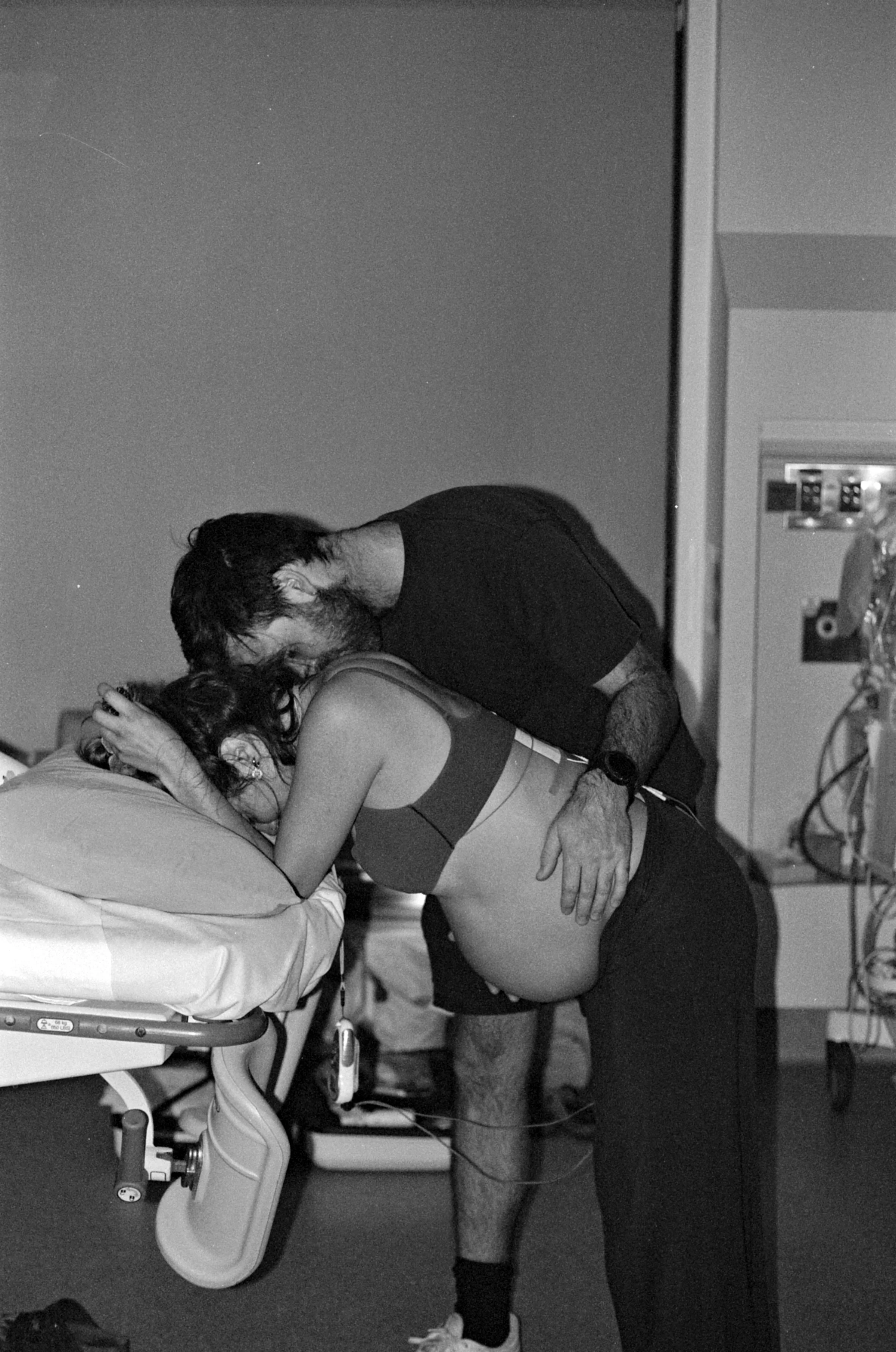 A couple sharing an intimate moment, with the man leaning over the woman, who is sitting on a hospital bed. The woman is wearing hospital attire, and the man is dressed casually. They are kissing, and the woman's hand is holding the man's head. The scene appears to be in a hospital room, with medical equipment and a bed visible in the background.
