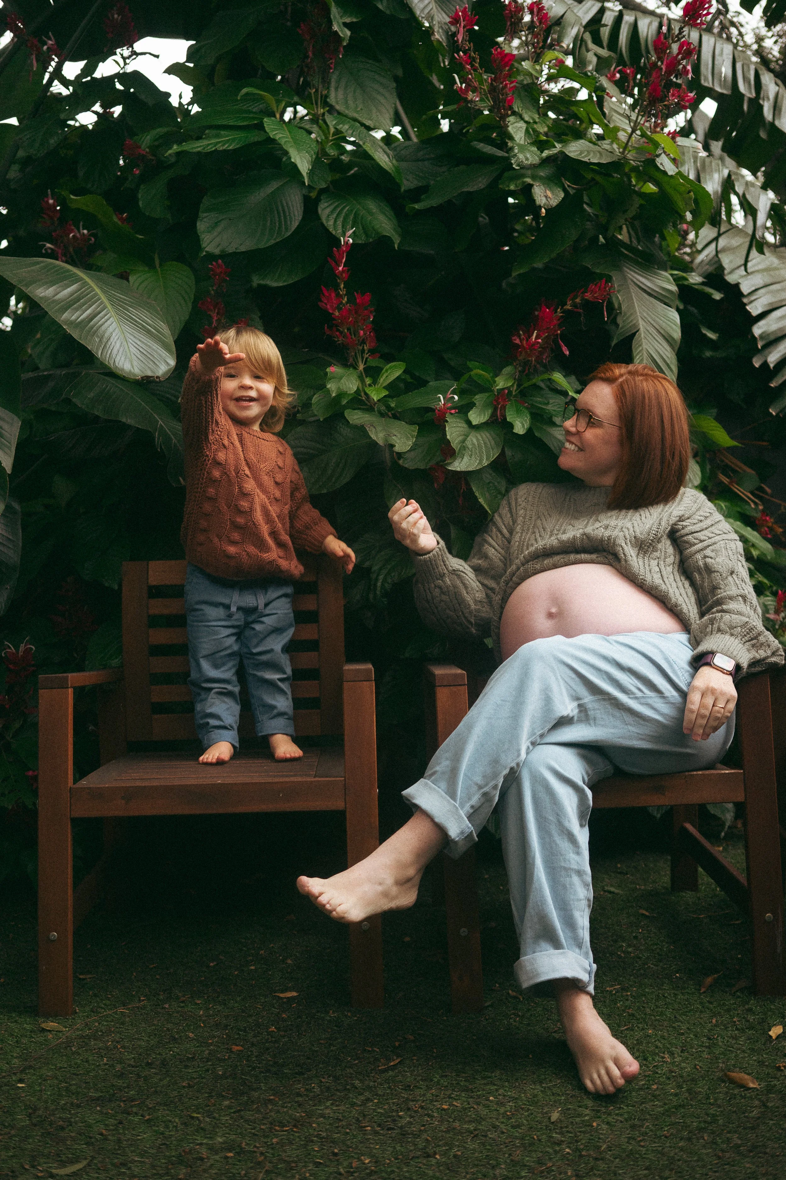 A woman with red hair and glasses sitting on a wooden chair with her pregnant belly visible, smiling at a young boy standing on a wooden platform. The boy has blonde hair and is waving and smiling, wearing a brown sweater and blue pants. They are outdoors surrounded by large green leaves and red flowers.