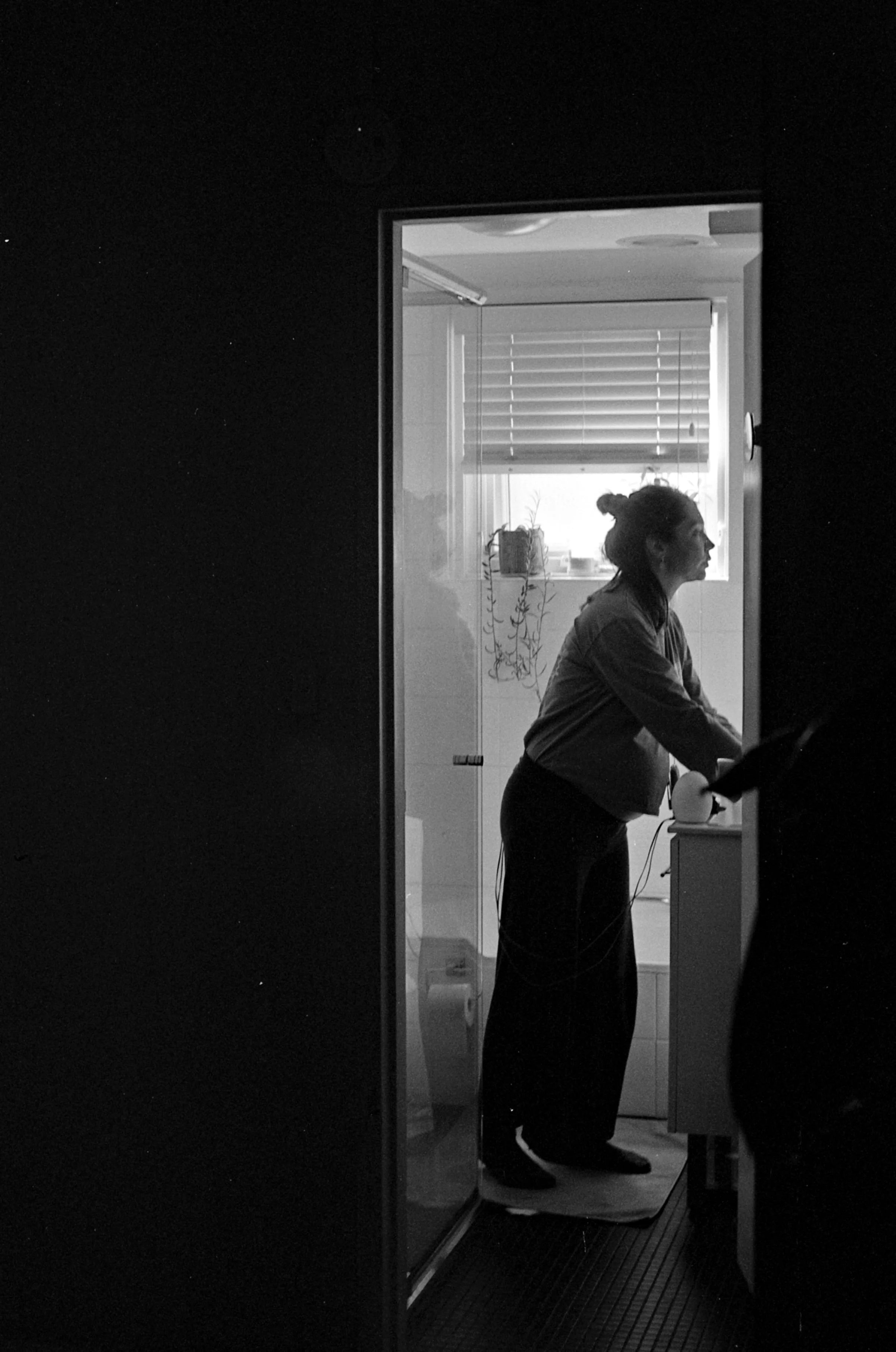 A black and white photo of a woman standing in a kitchen, seen from a side profile, through an open doorway, illuminated by sunlight from a window with blinds.