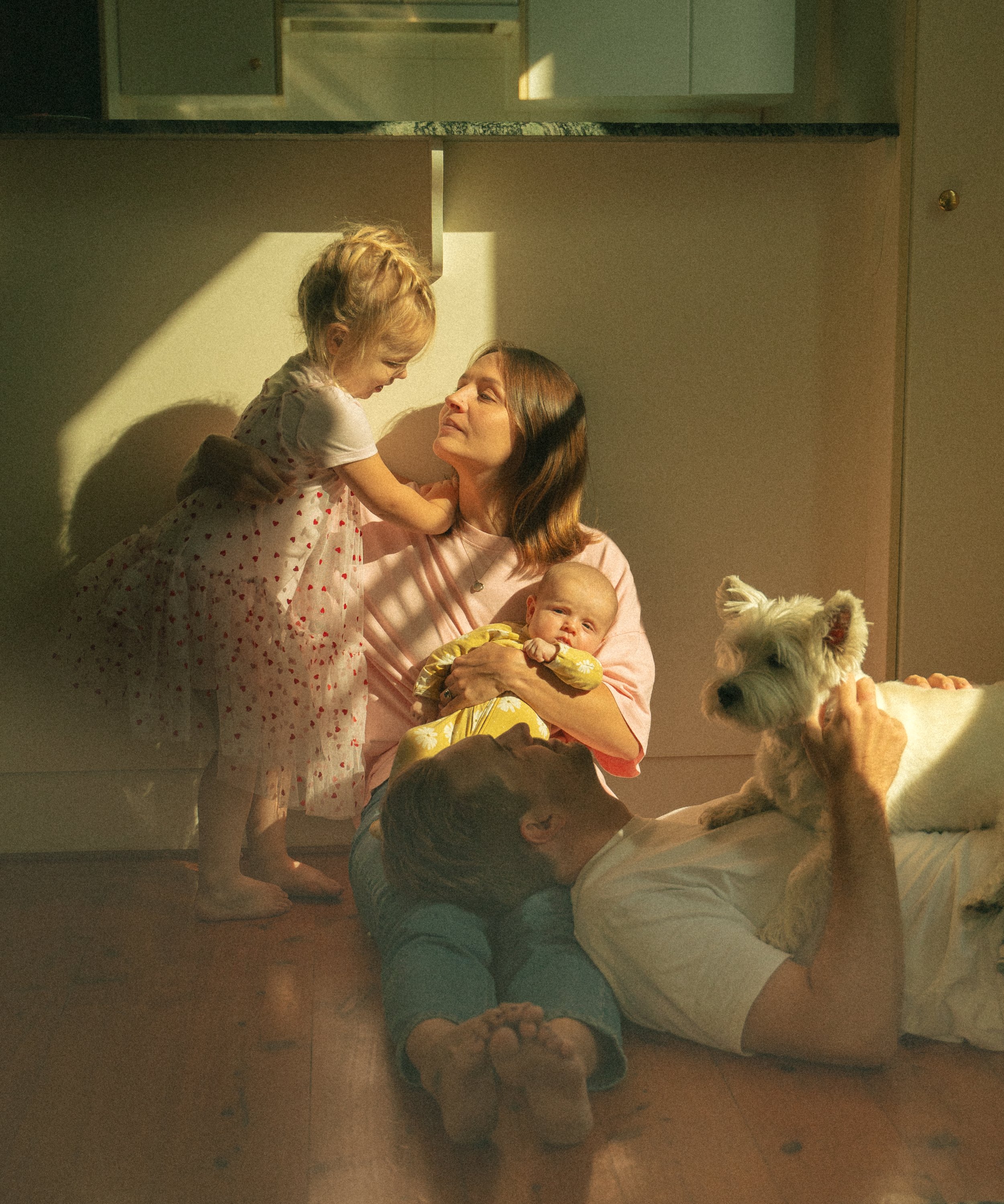 A multi-generational family in a cozy room with sunlight, including a woman lying on the floor with a dog on her stomach, a young girl standing and leaning in, another woman sitting on the floor holding a baby, and a young boy lying on the floor.