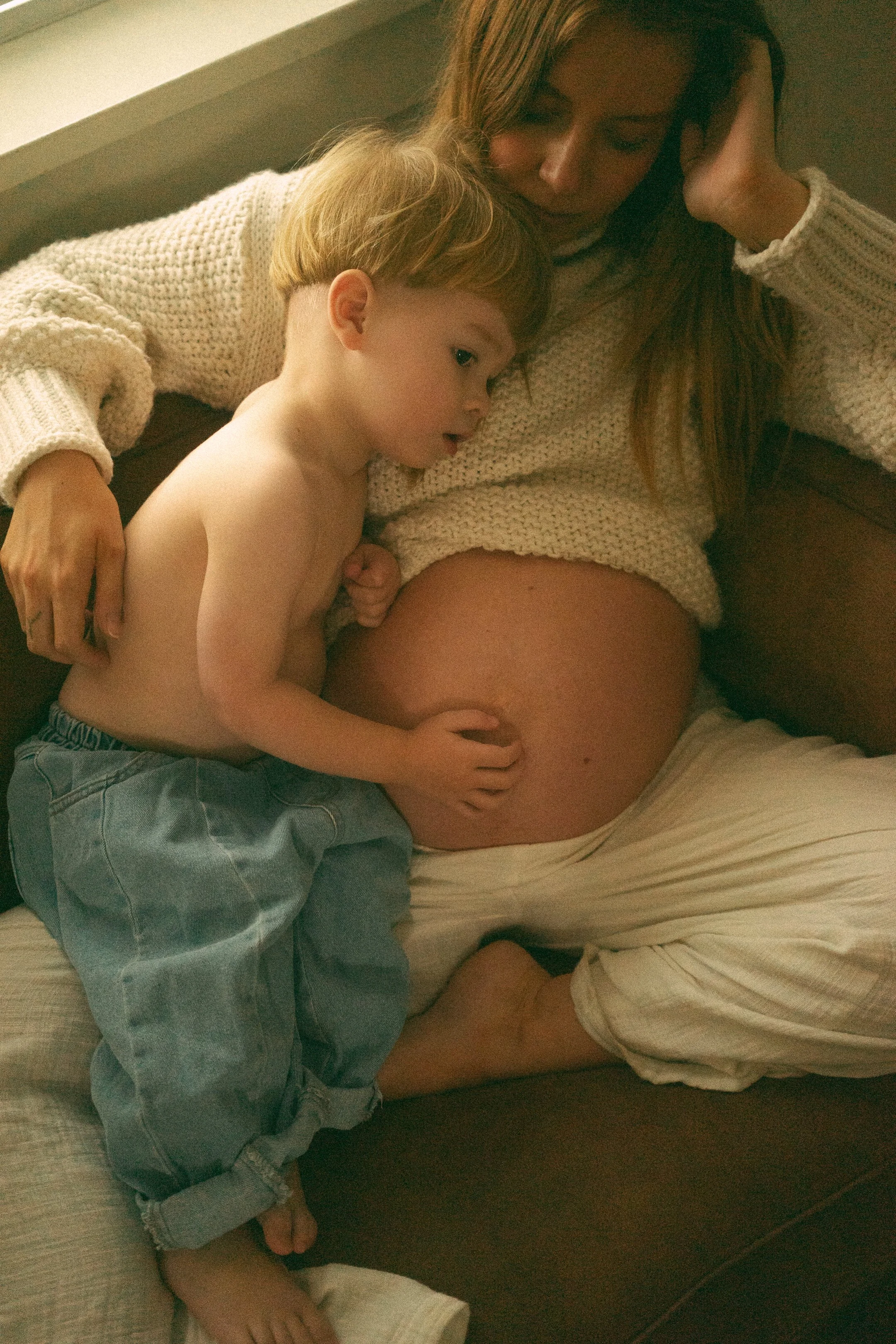 A woman with long hair in a white sweater lying on a couch, with a young child abdomen and chest resting on her, looking at her belly.