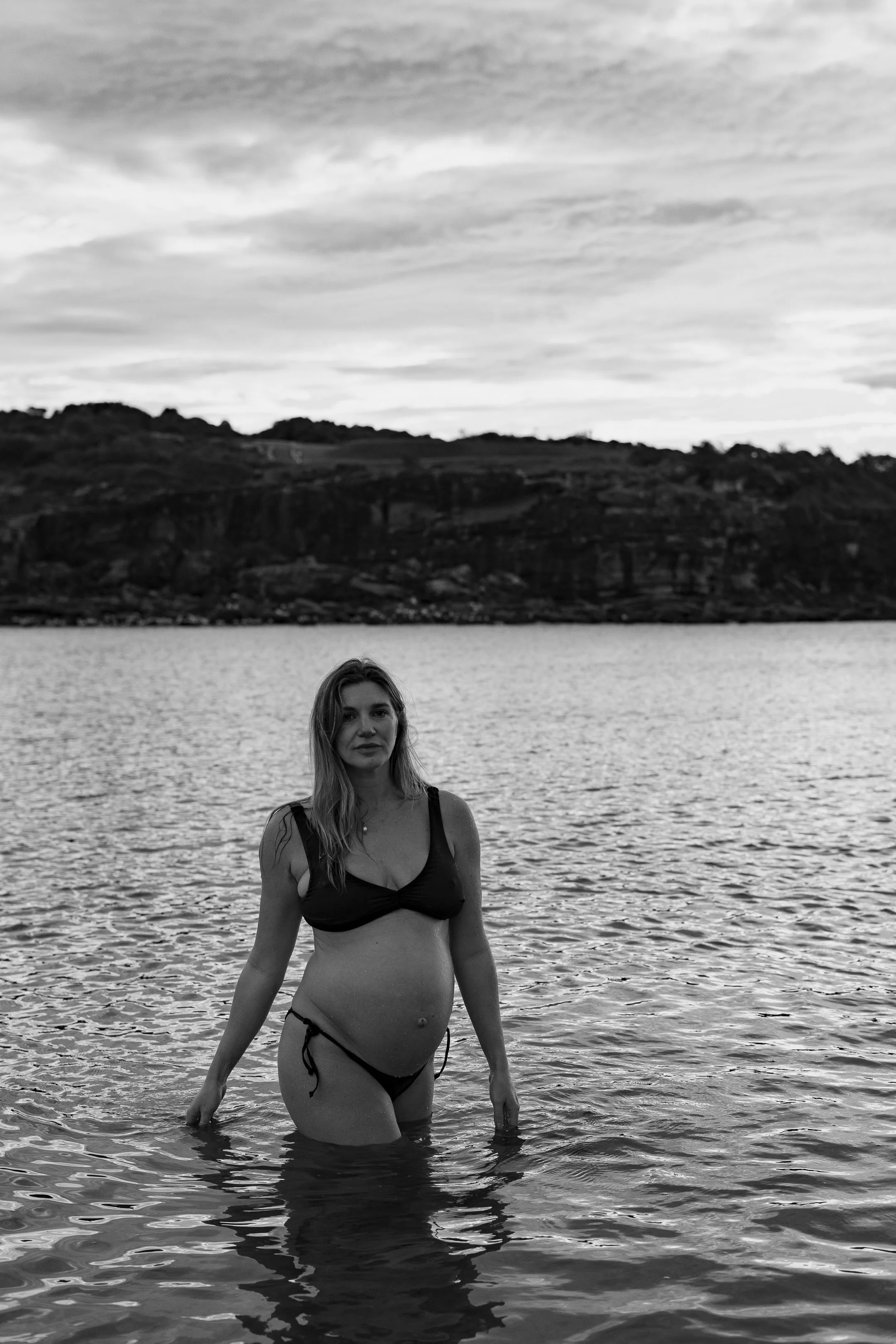 A woman standing in water near a shoreline with hills and cloudy sky in the background. She is wearing a bikini and looking at the camera.