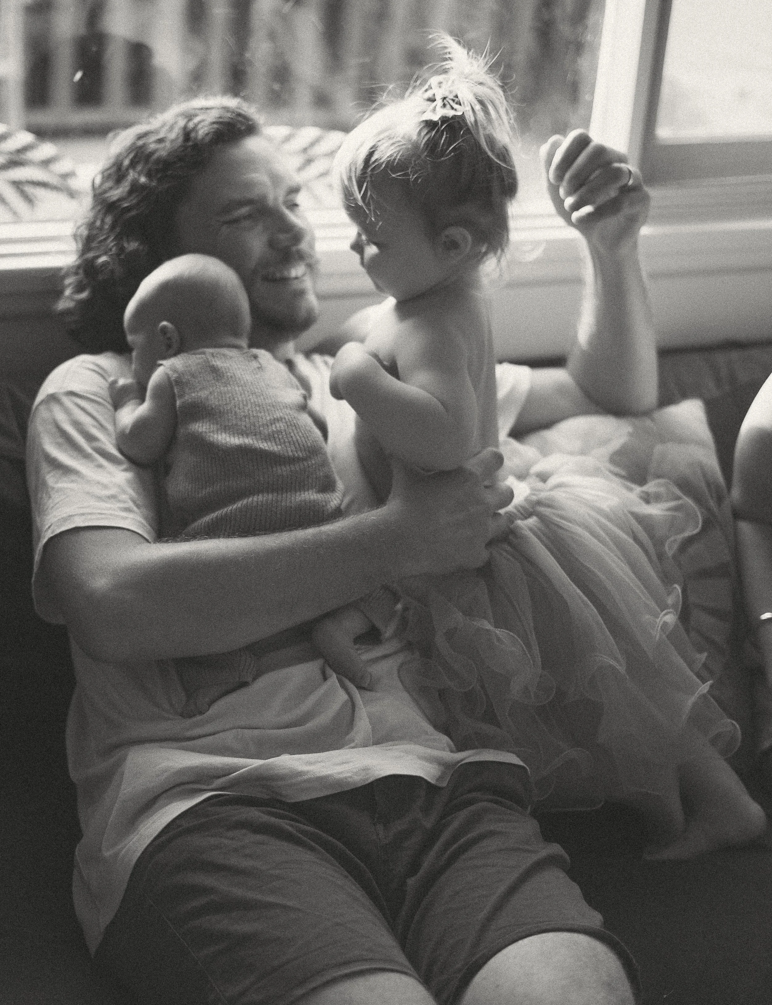 A man with curly hair and a beard sitting on a couch near a window, holding two young children. One child is a baby pressed against his chest, and the other is a toddler girl with tousled hair, wearing a tutu skirt, sitting on his lap and touching his face. They are smiling and enjoying a tender moment together.