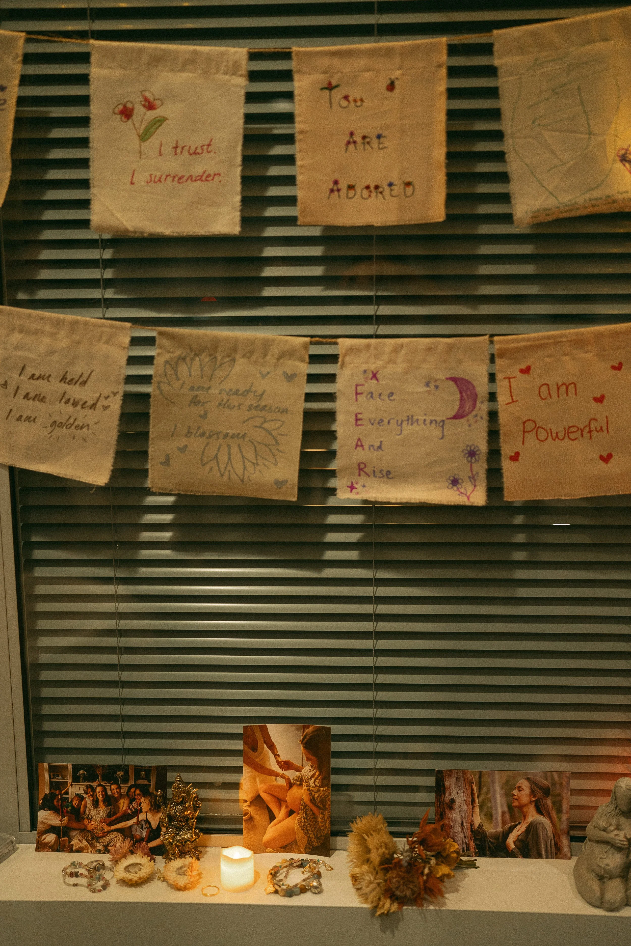 Display of handmade fabric banners with inspirational messages and drawings hanging on a window blind. Below the banners is a table with photographs, flower arrangements, jewelry, and a lit candle.