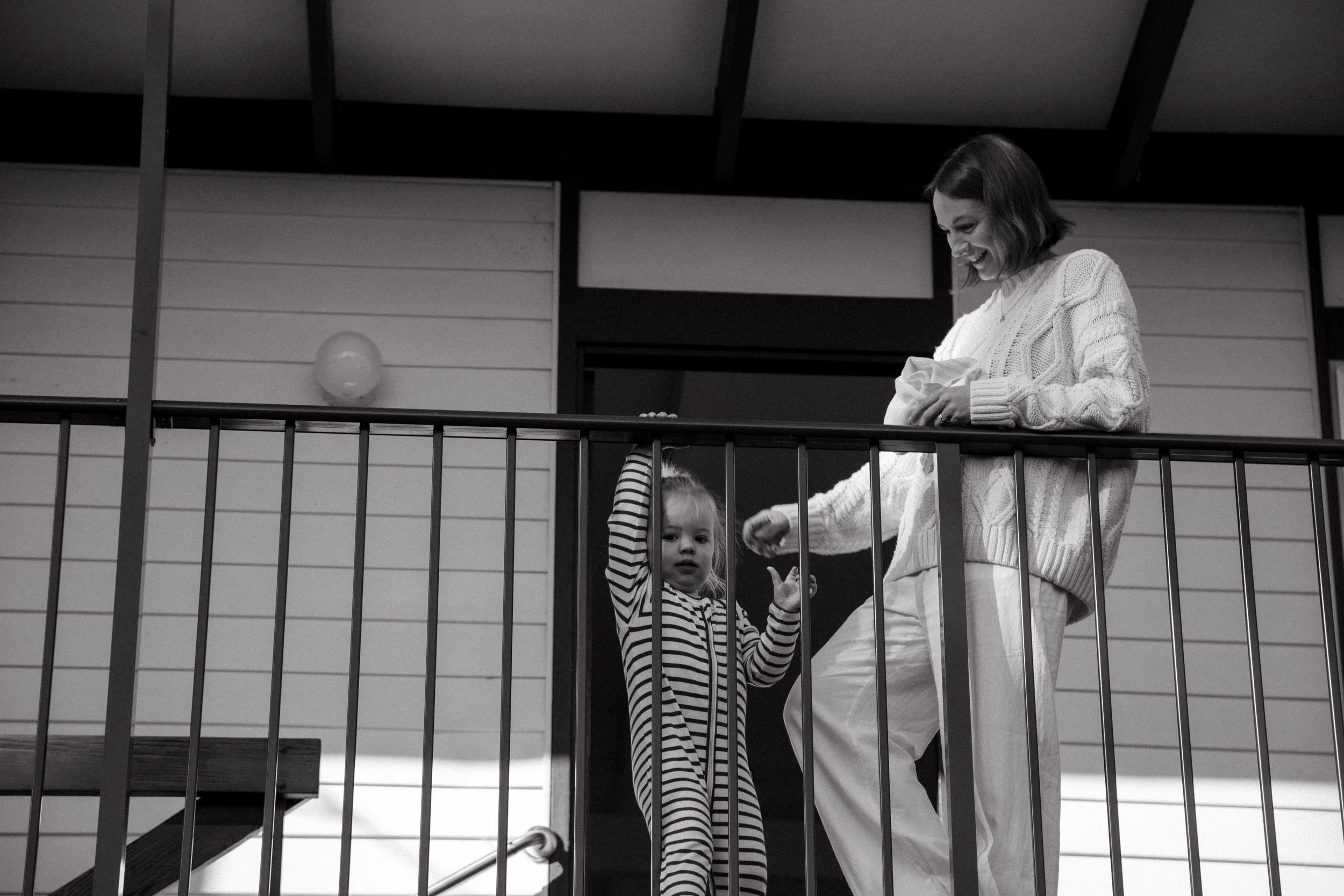 A woman and a young girl on a balcony. The woman is smiling and handing something to the girl who is wearing a striped onesie and looking at the camera.