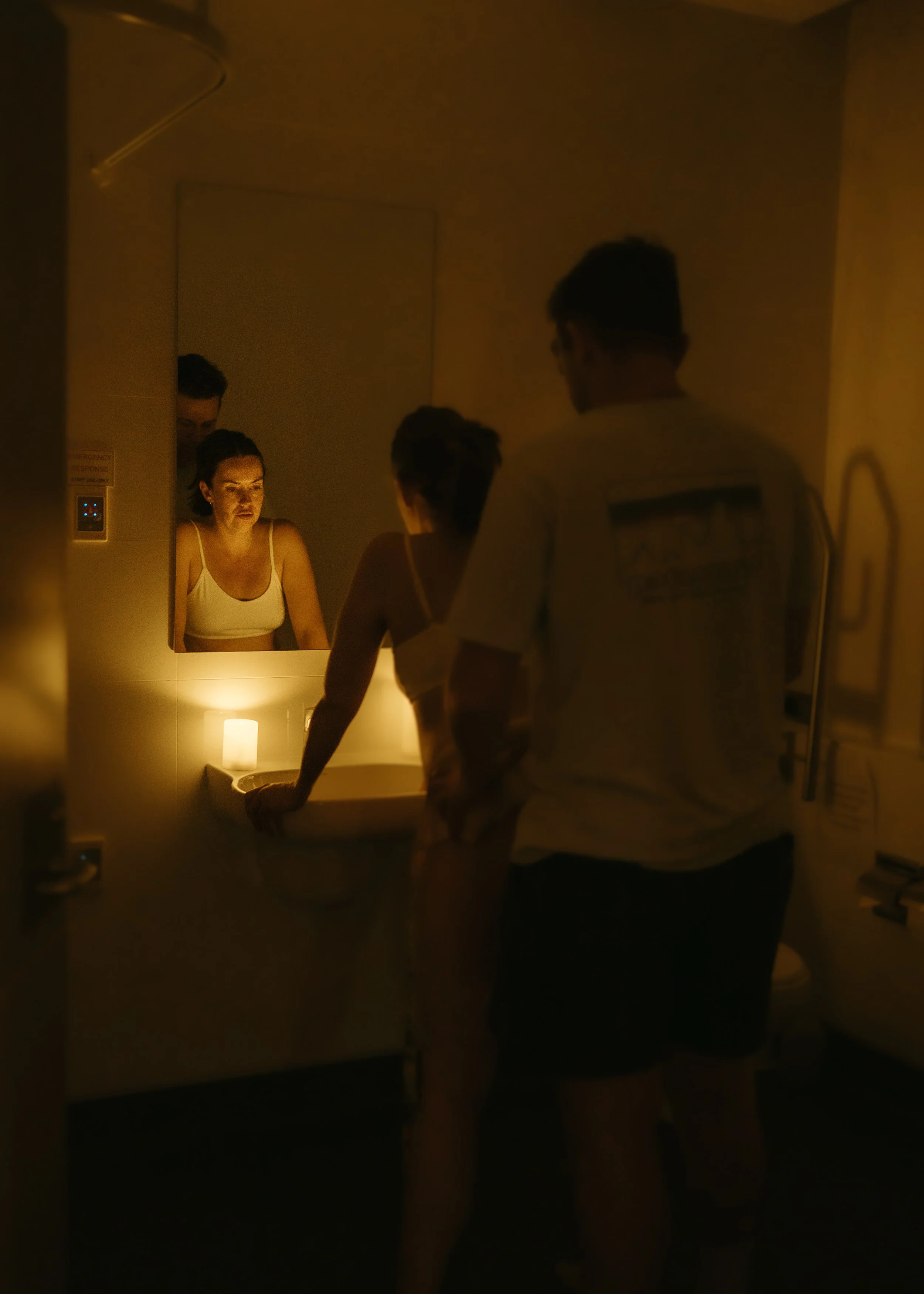 A woman is in a dimly lit room, standing at a sink in front of a mirror, with her reflection visible. A man stands nearby, possibly talking to her, and a small candle provides warm light in the scene.