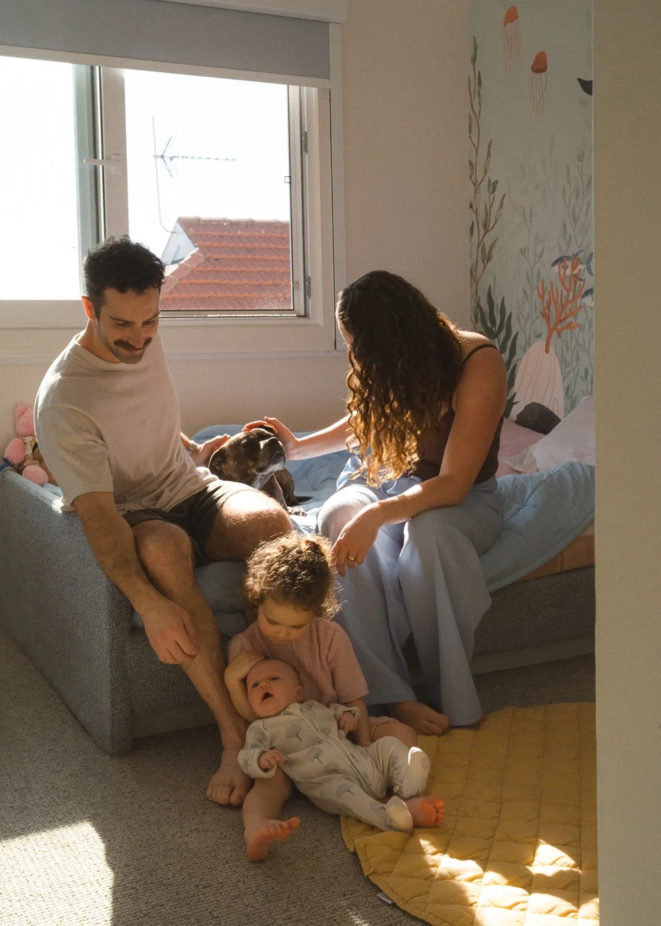 A family of four, including two children and a dog, sitting on and around a bed in a sunlit bedroom with a mural of jellyfish and sea plants on the wall.