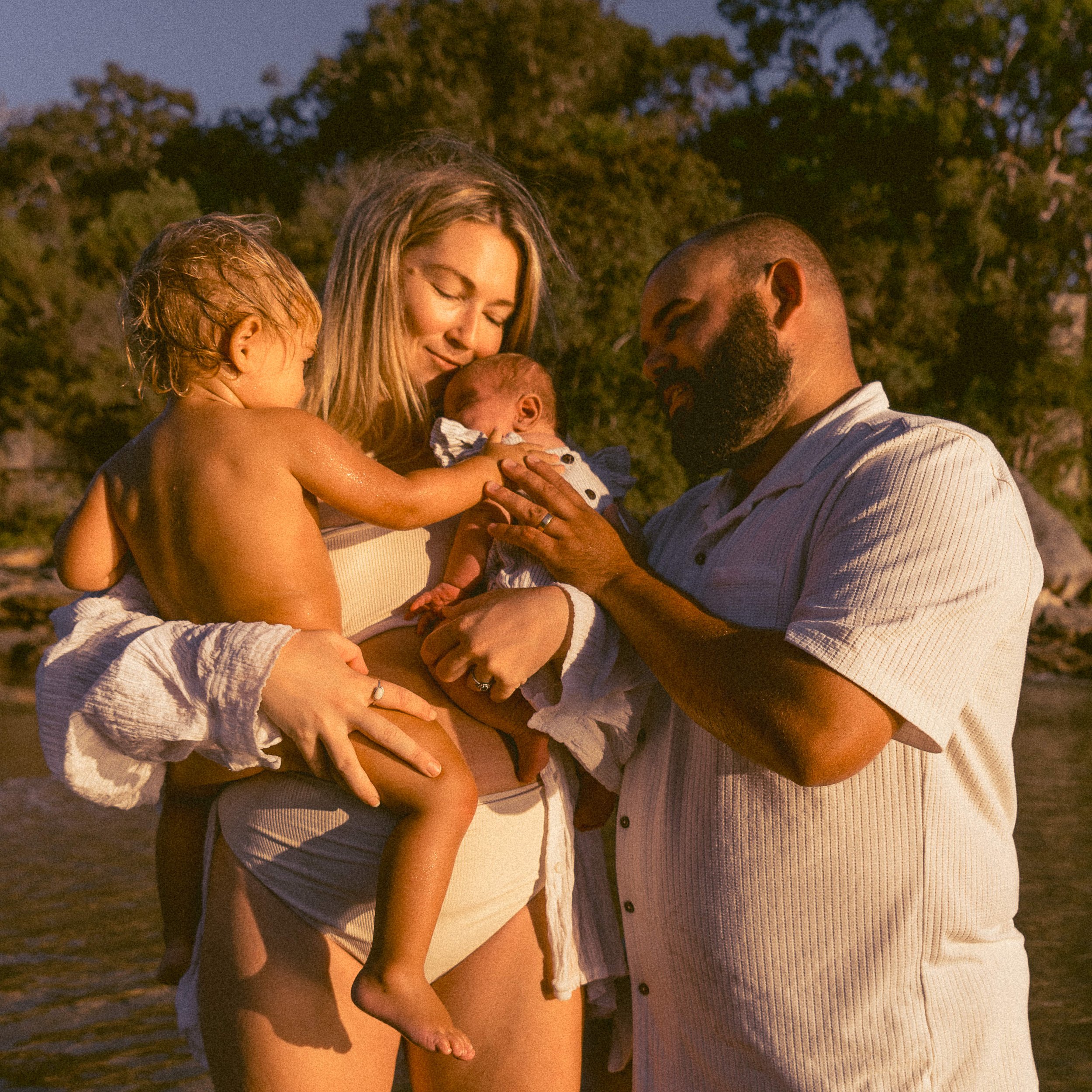 A family of four, including a woman, a man, a young child, and a newborn baby, enjoying a moment together outdoors near water during sunset.