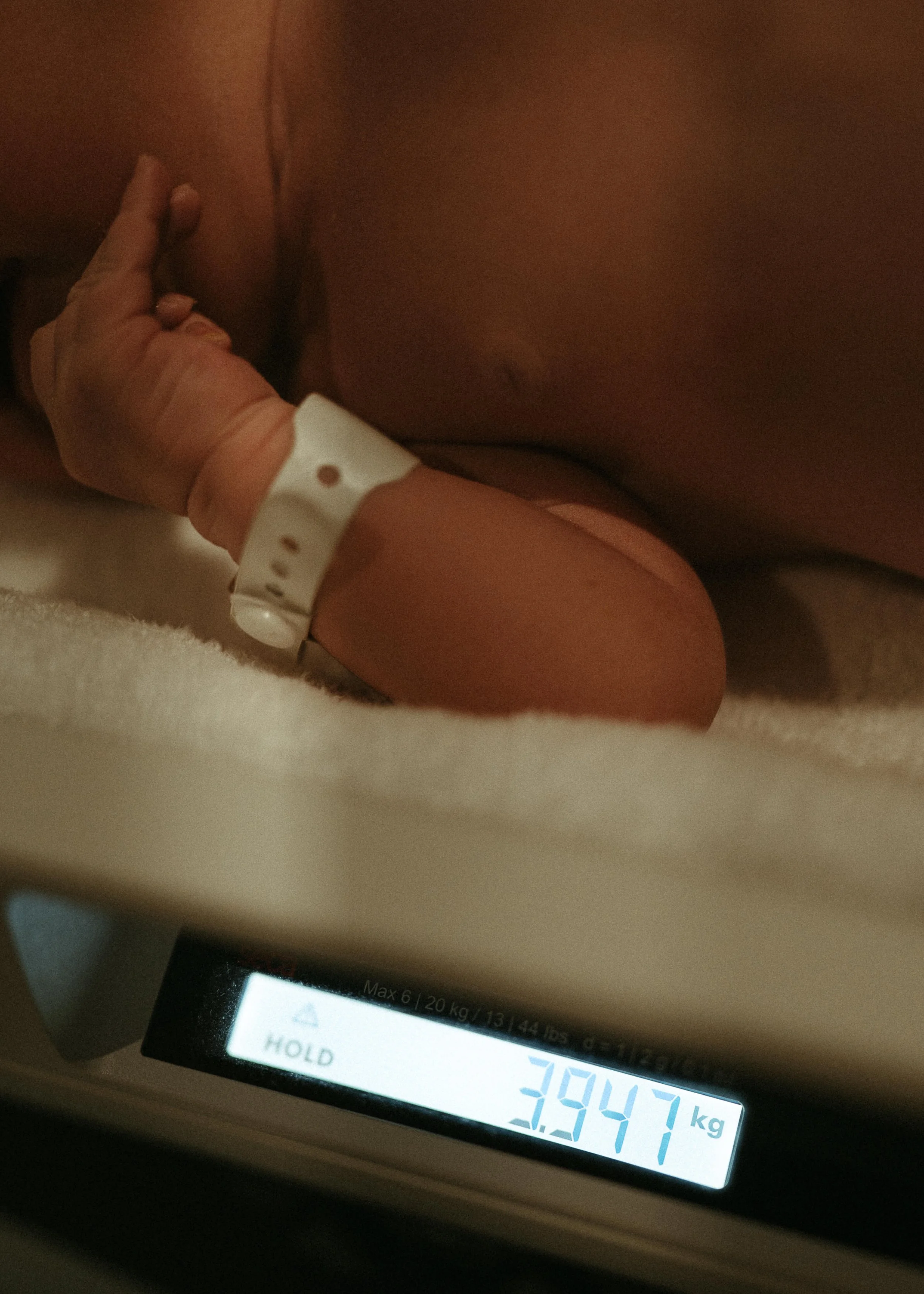 A person’s hand with an ID bracelet resting on their knee near a digital scale displaying a weight of 3.947 kilograms, with part of the display screen visible showing text and numbers.