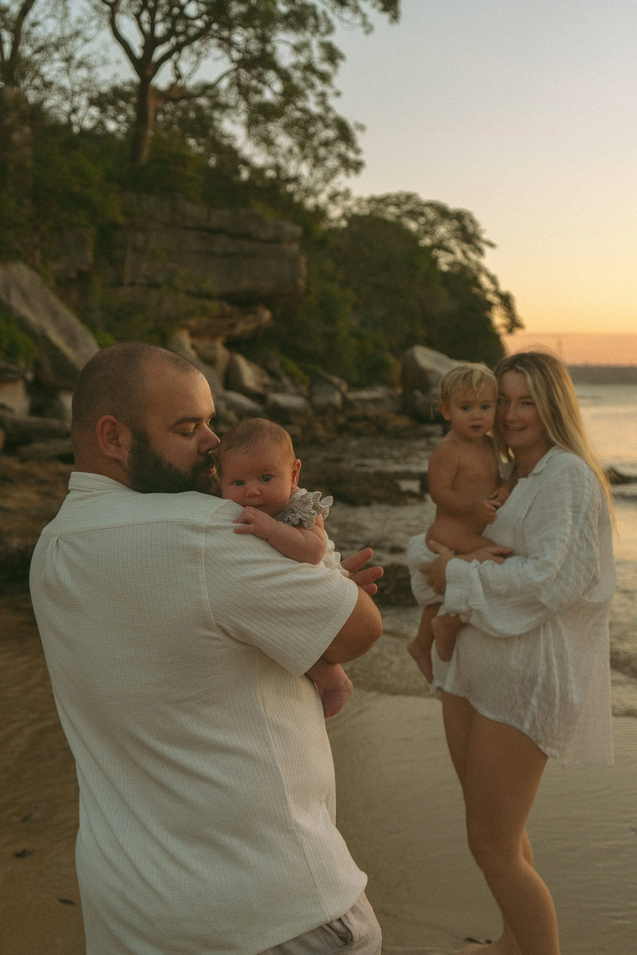 A family of four with two young children enjoying a sunset on the beach, with trees and rocks in the background.