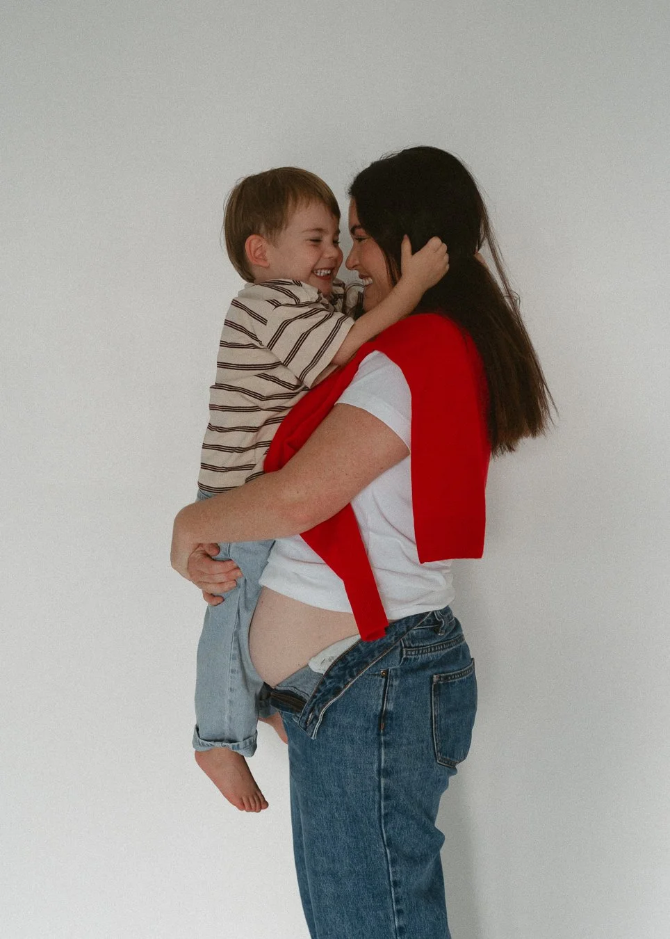 A woman is holding a young boy, both smiling and touching foreheads, against a plain white wall.