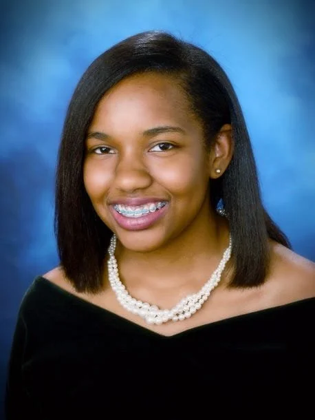 A young woman with straight black hair, smiling and showing braces, wearing a pearl necklace and a black off-the-shoulder top, posed against a blue background.