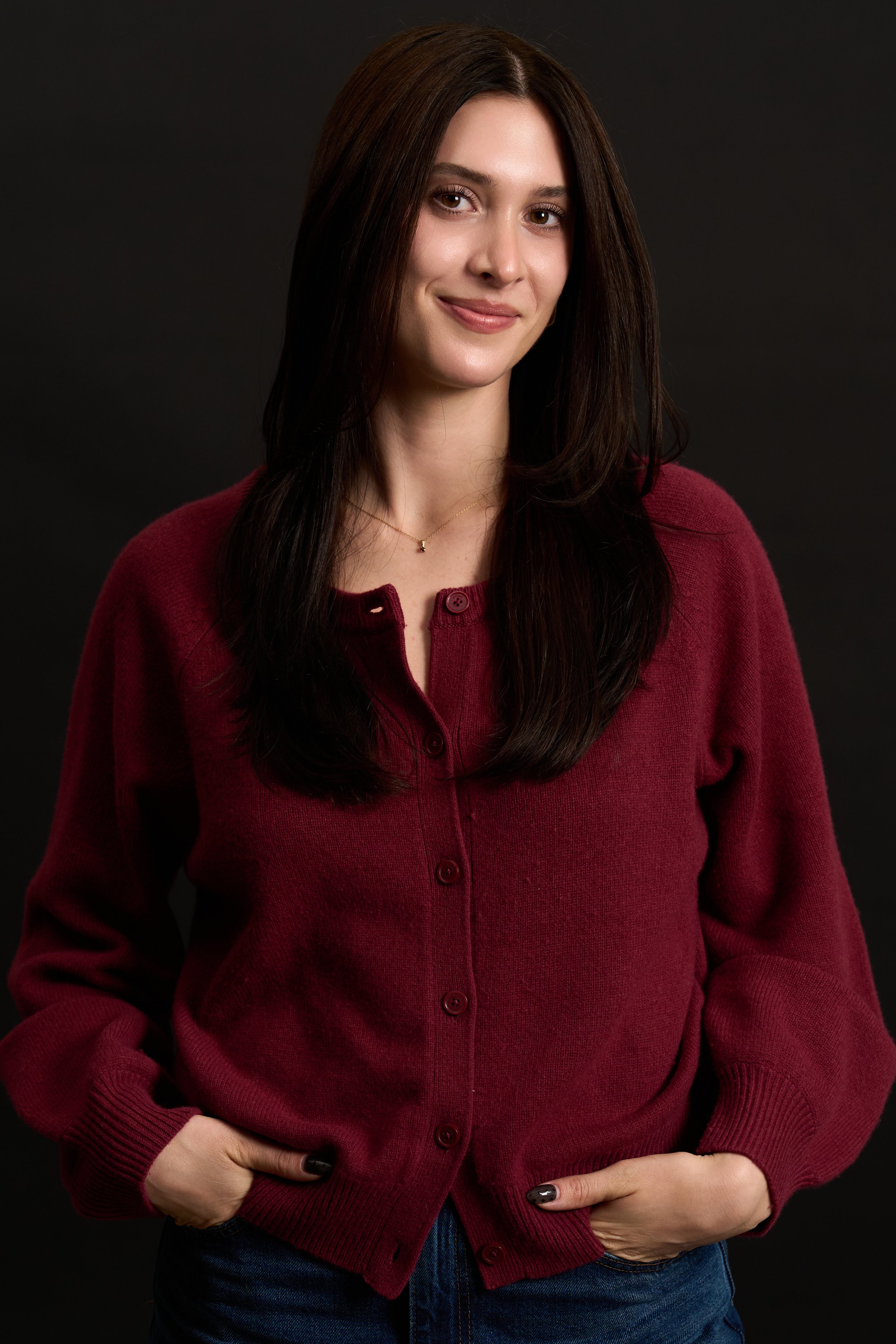 A young woman with long dark hair wearing a burgundy cardigan and blue jeans, standing against a black background, smiling slightly, with her hands resting in her pockets.