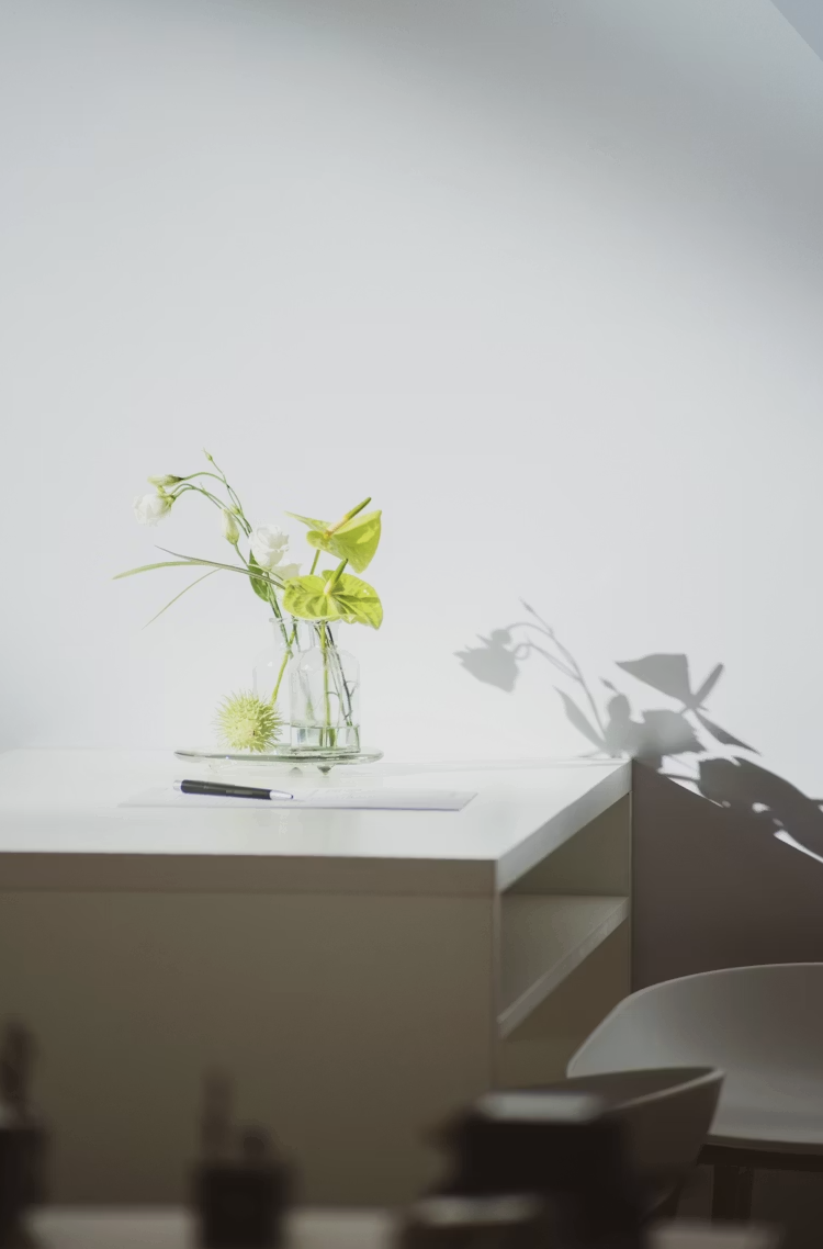 A minimalist white desk with a glass vase containing green and white flowers and leaves, casting a shadow on the wall behind. A black pen and a piece of paper lie on the desk, with a white chair partially visible in the foreground.