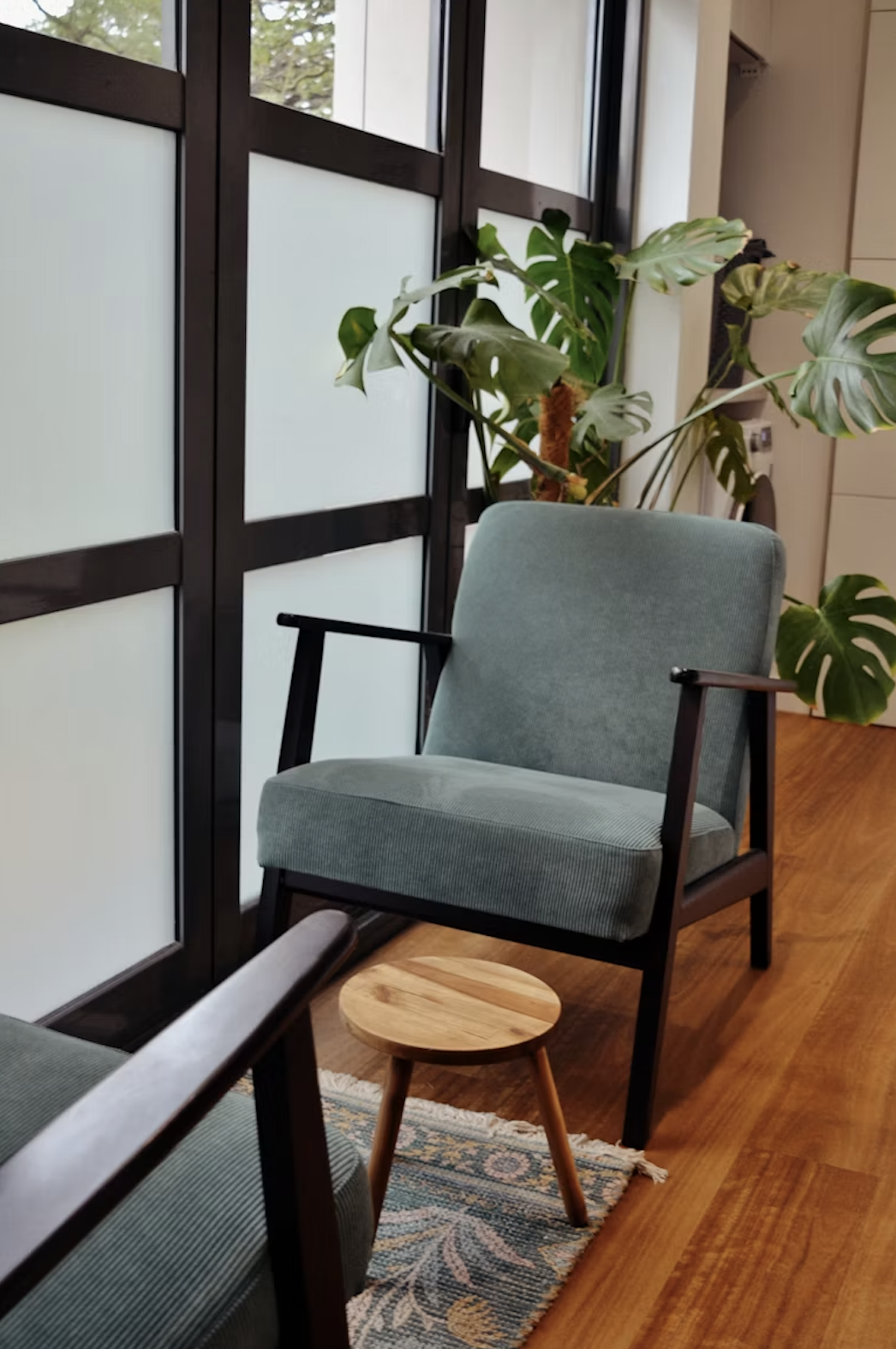 A cozy indoor space with a light green armchair, a small round wooden table, and large leafy houseplants near a window with frosted glass panels.