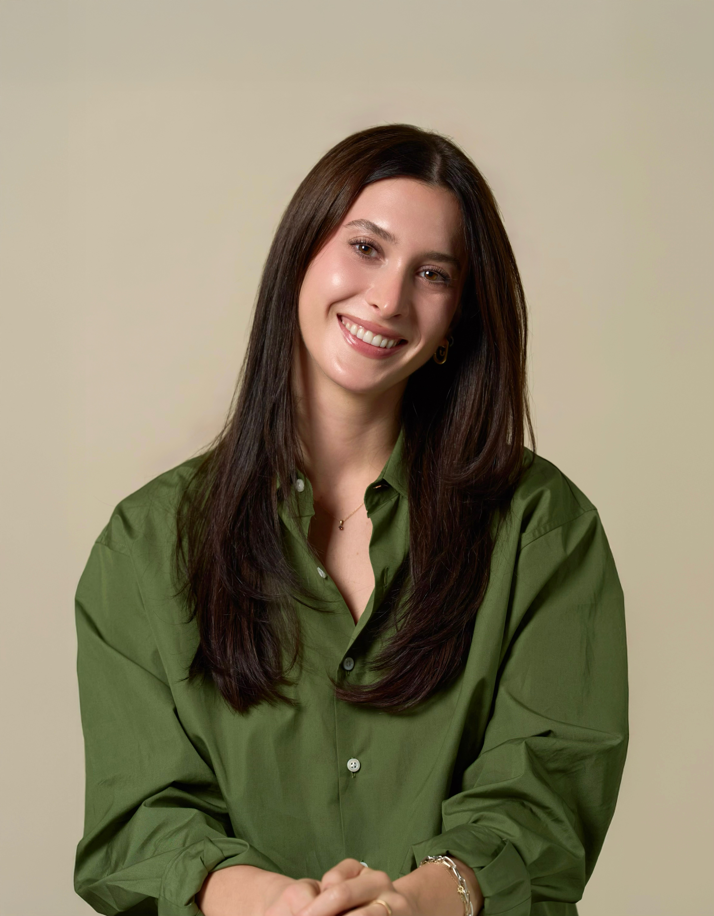 A woman therapist with long dark brown hair, smiling, wearing an olive green shirt, gold earrings, and a gold bracelet, sitting against a plain light beige background.