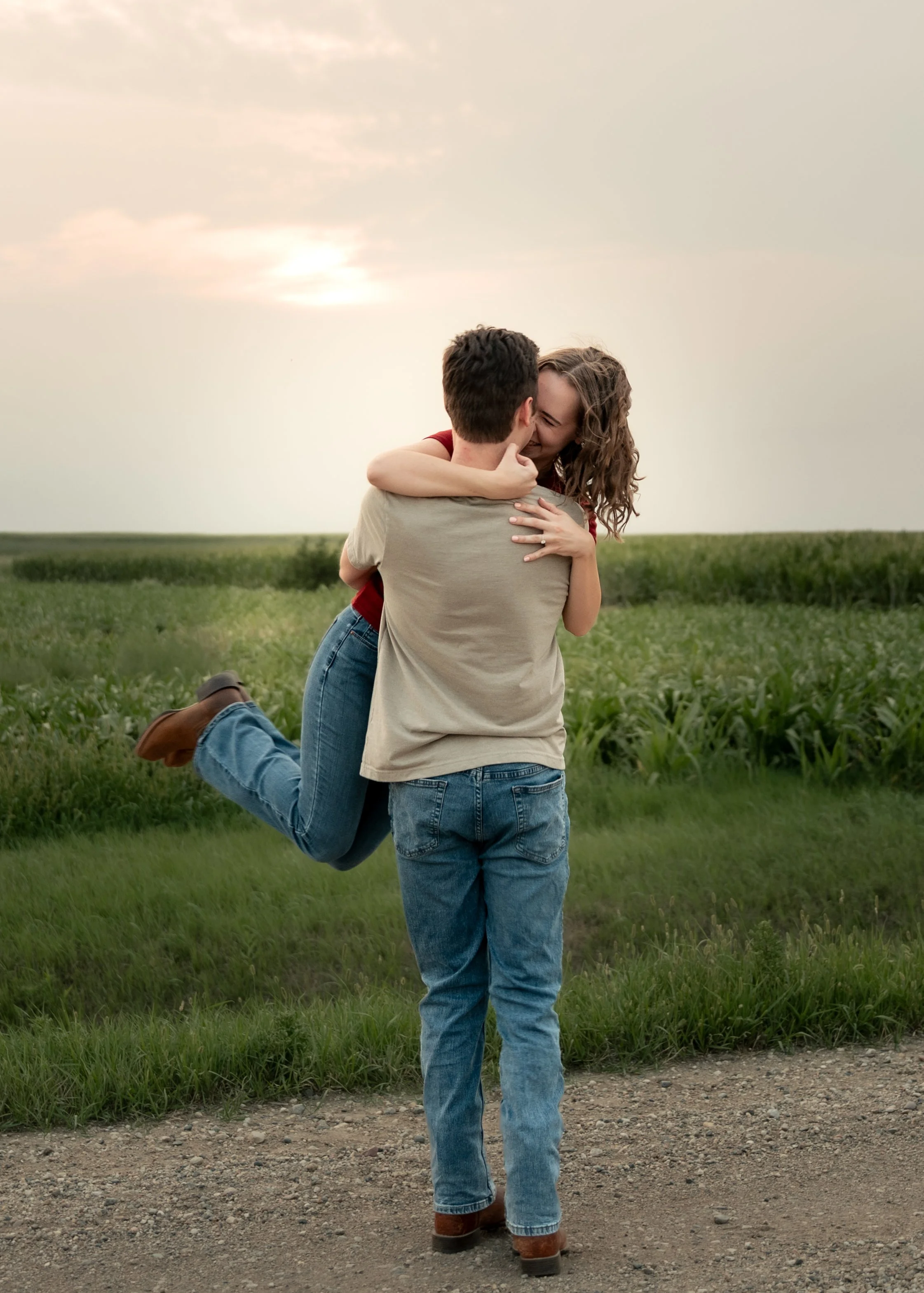 A couple dressed in casual clothing embrace outdoors on a dirt path, with the woman being lifted off the ground while smiling and the man facing away, in a grassy field with a cloudy sky in the background.