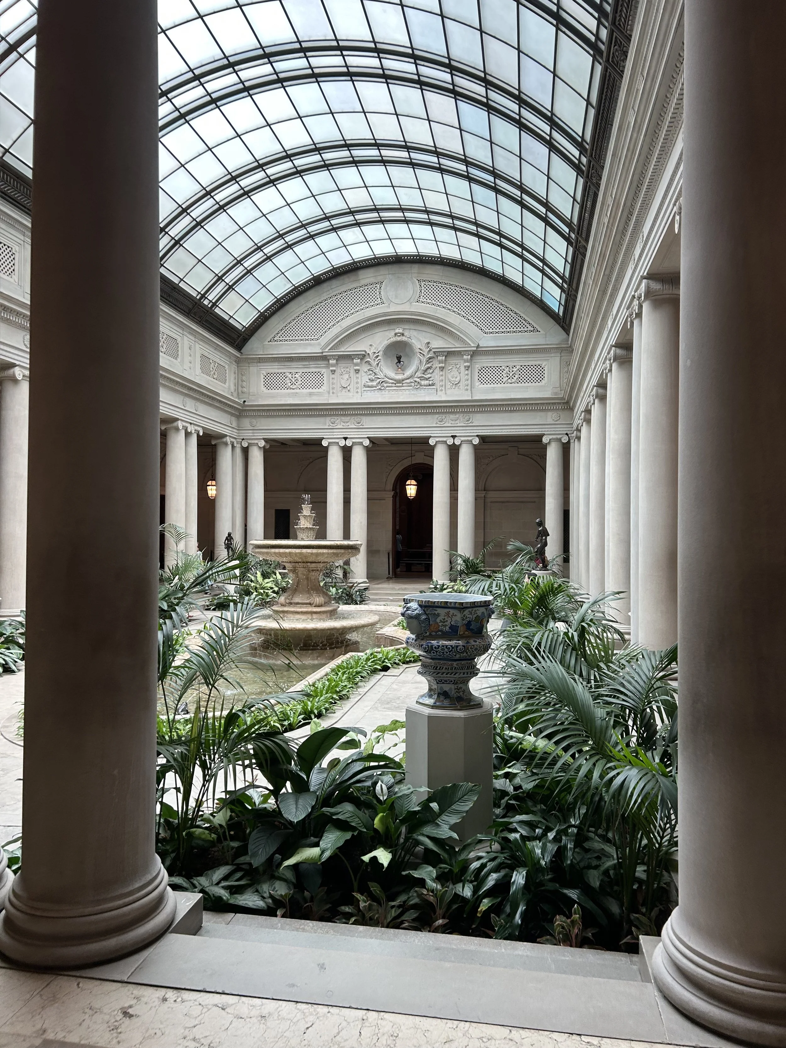 An elegant indoor courtyard with a glass-domed ceiling, classical white columns, decorative moldings, a fountain, various statues, lush green plants, and ornate vases on pedestals.