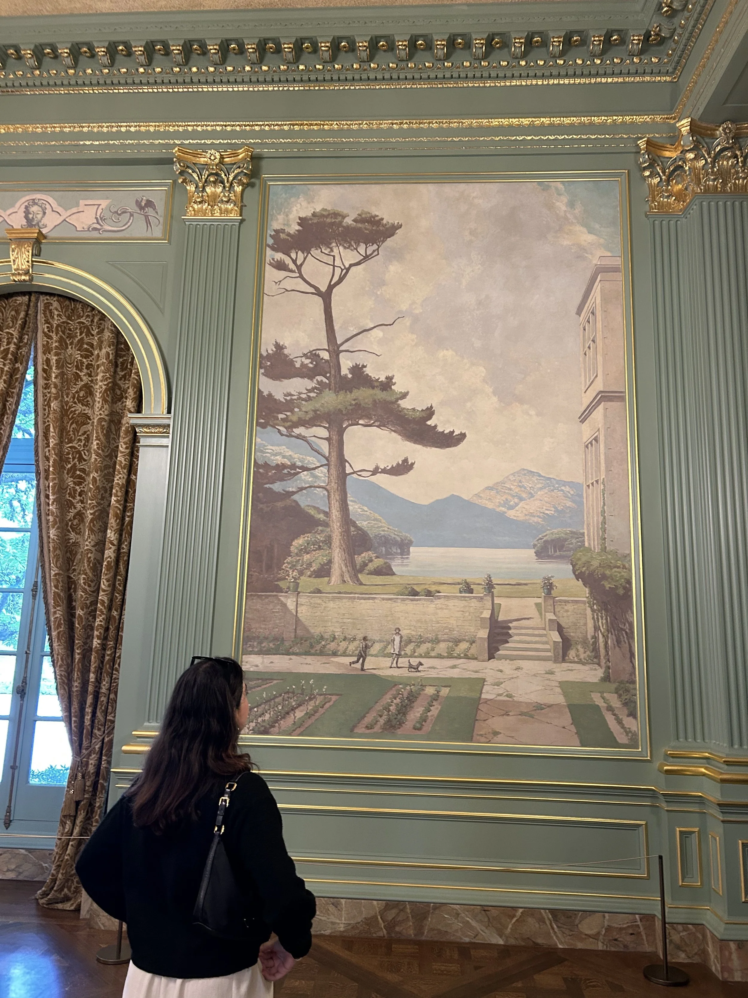 A large landscape mural showing a tall pine tree by a river, with mountains in the background. There are people walking and children playing in front of the mural. The room has ornate green and gold trim on the walls and ceiling, with gold accents an