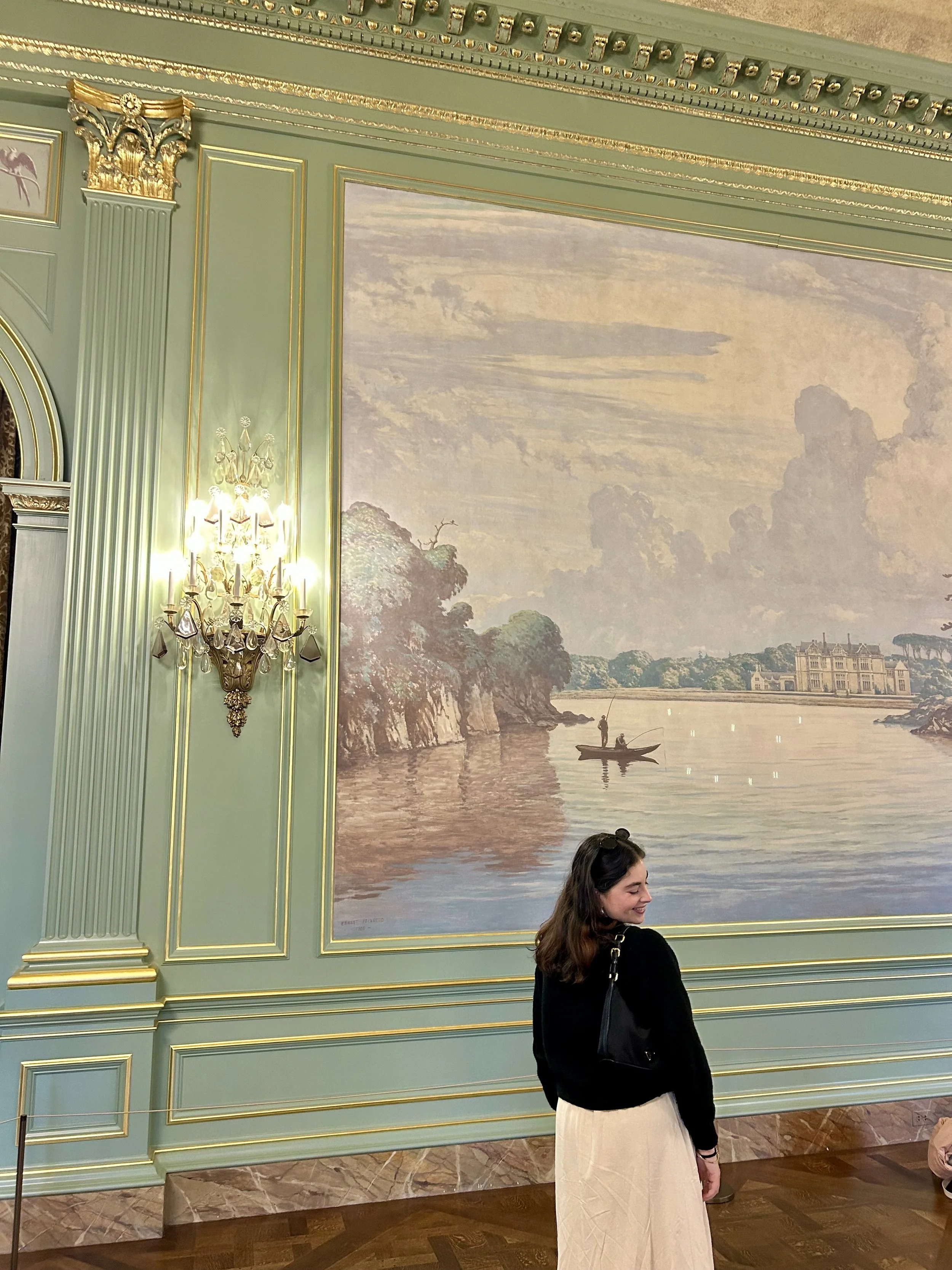 A woman with long brown hair wearing a black top and beige skirt is standing in front of an ornate wall with a large landscape painting. The wall is painted in pastel green with gold trim and detailed moldings. There is a chandelier with crystals and