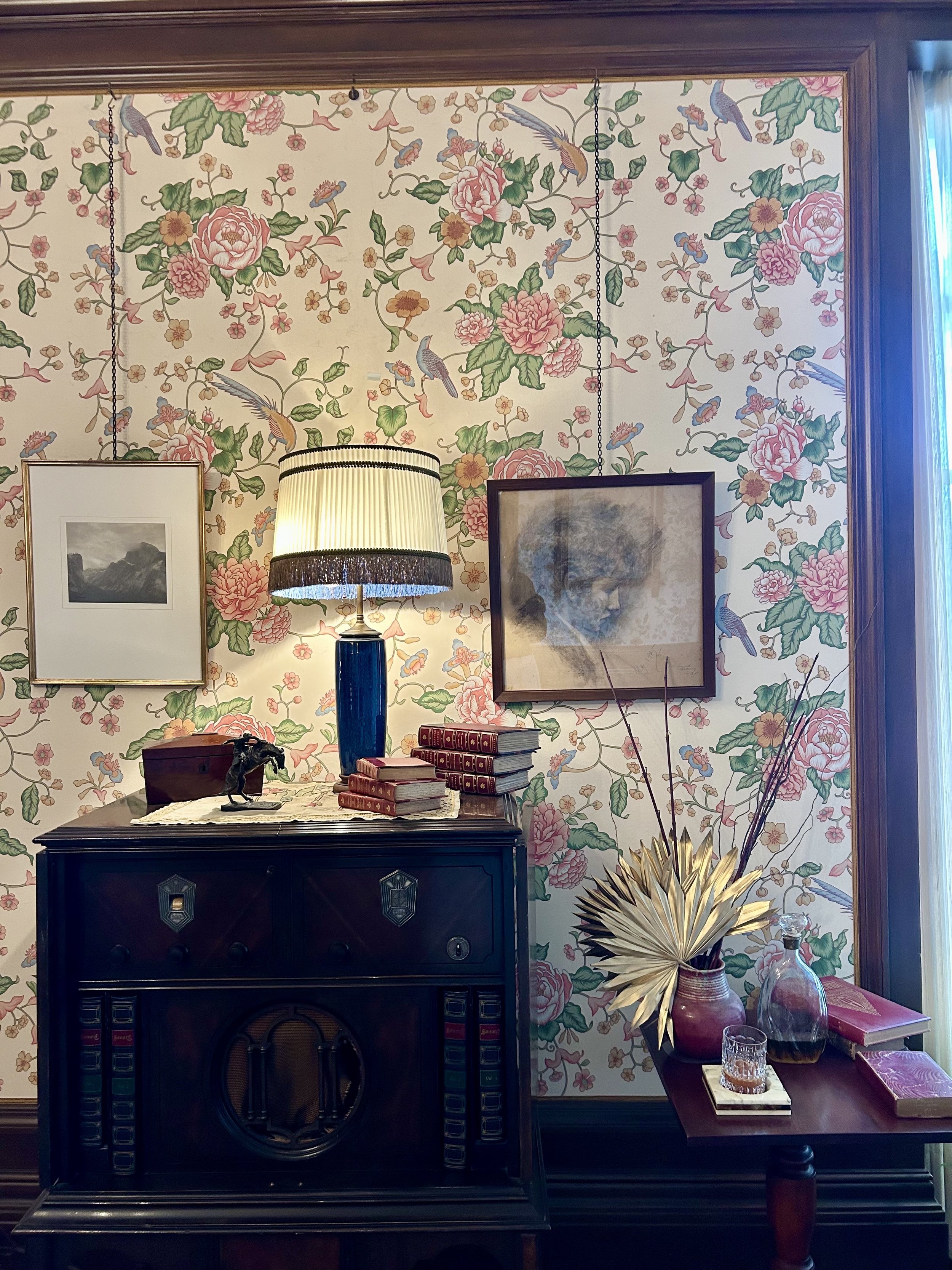 A vintage interior with floral wallpaper, a dark wood fireplace mantel, a table with stacked red books, a table lamp, and decorating objects including a sculpture, dried plant arrangement, a glass bottle, and assorted books.