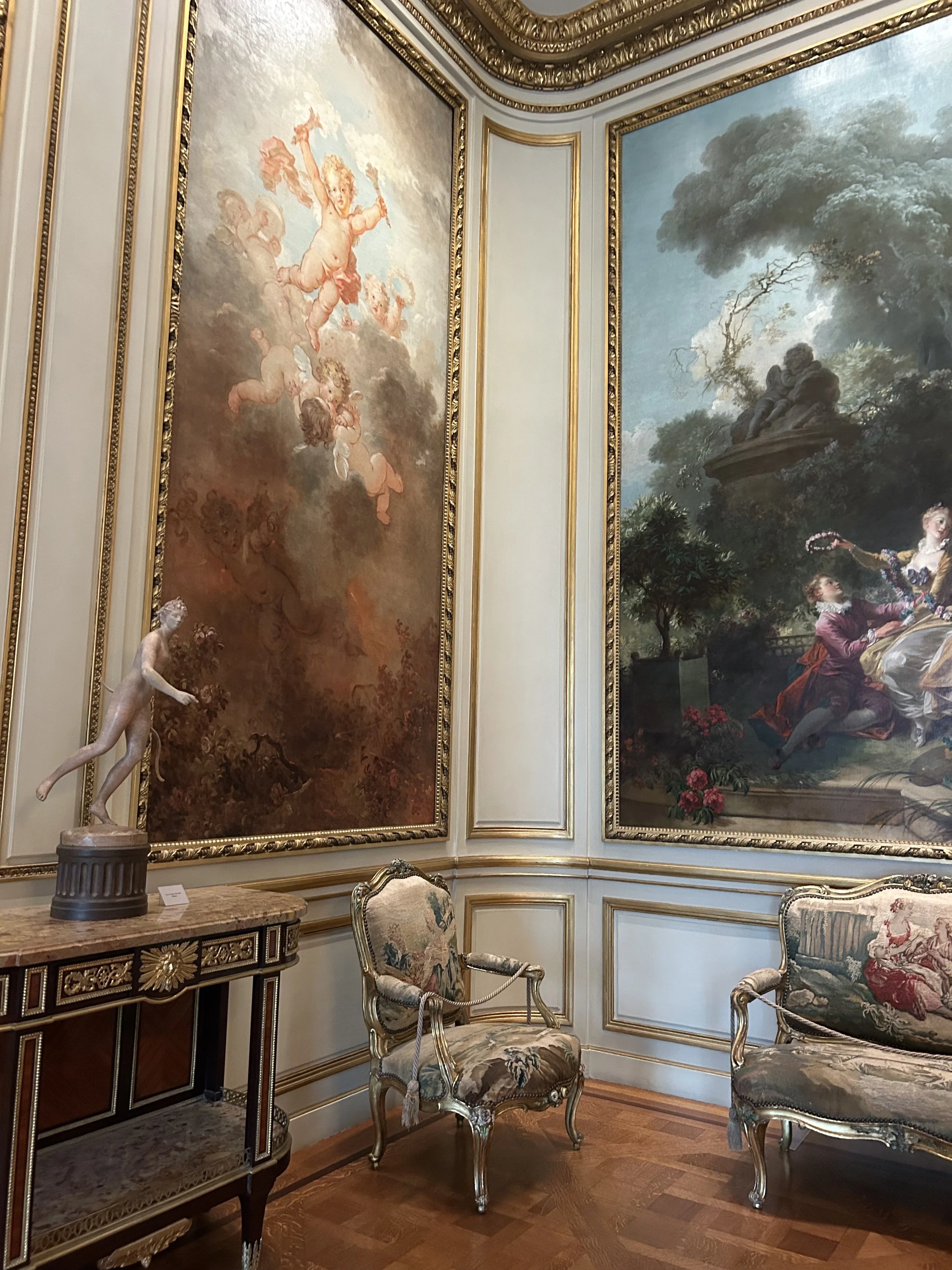 Elegant room with ornate wall moldings and large framed paintings. There is a marble-topped console table holding a classical-style sculpture of a standing female figure, and two upholstered chairs with carved wood frames and floral fabric. The hardw