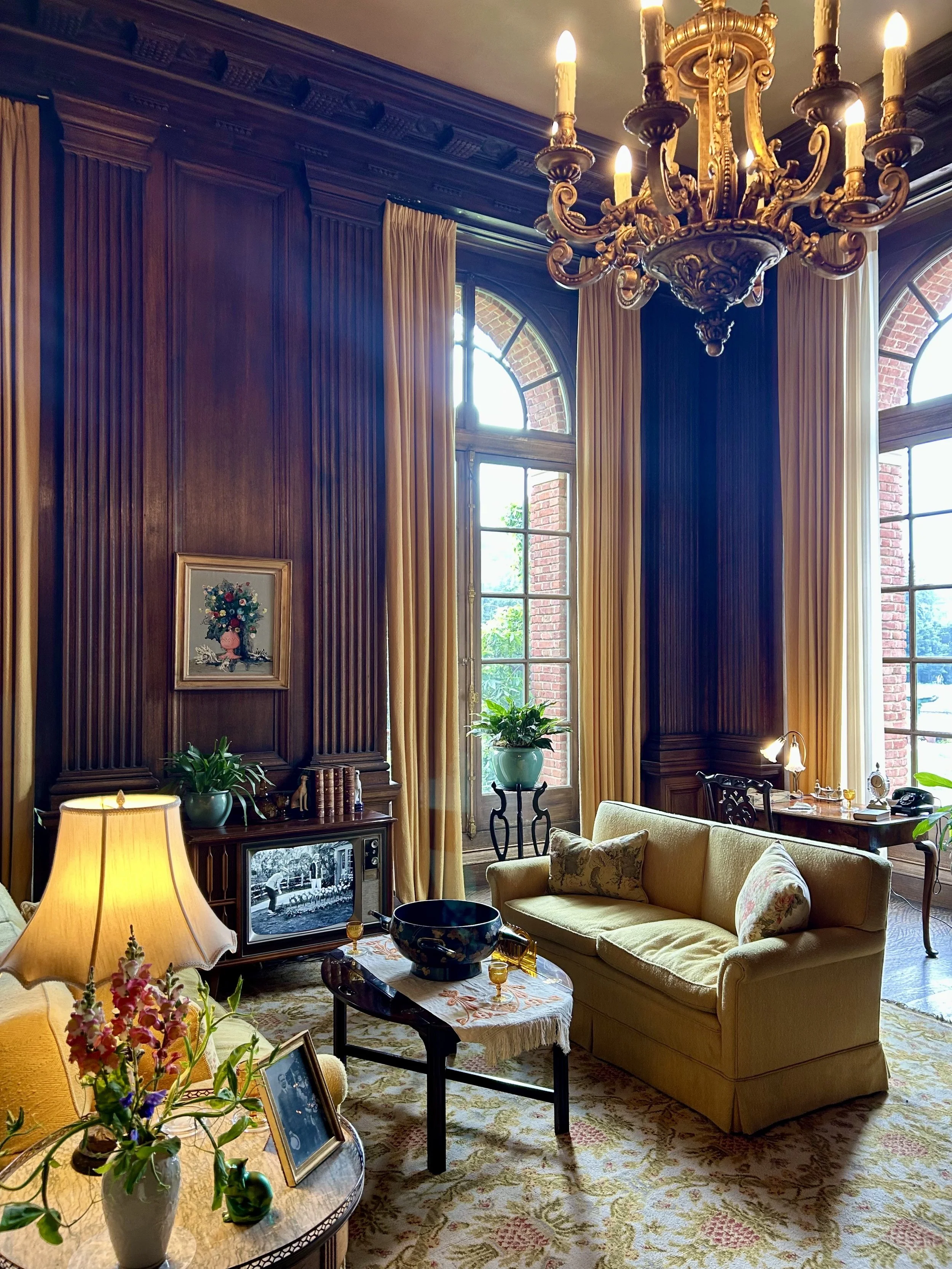 A living room with wooden paneling, tall arched windows with yellow curtains, a chandelier, a sofa with floral pillows, a table with a tablecloth, a television, and various decorative items.