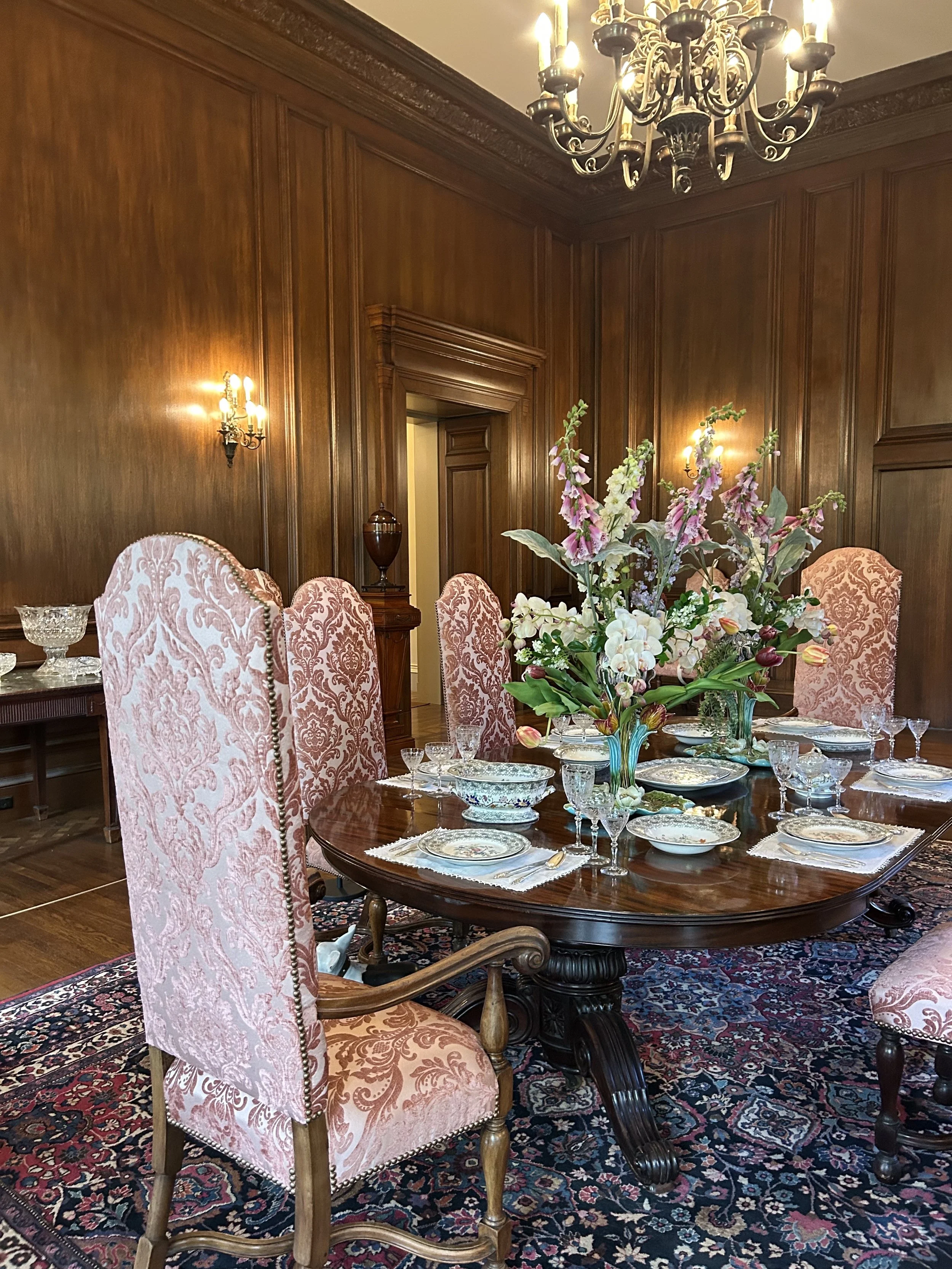 A formal dining room with a polished wooden round table set with fine china, glassware, and silverware, and a large floral centerpiece. The room features pink upholstered high-back chairs with intricate patterns, wood-paneled walls, a Persian rug, an