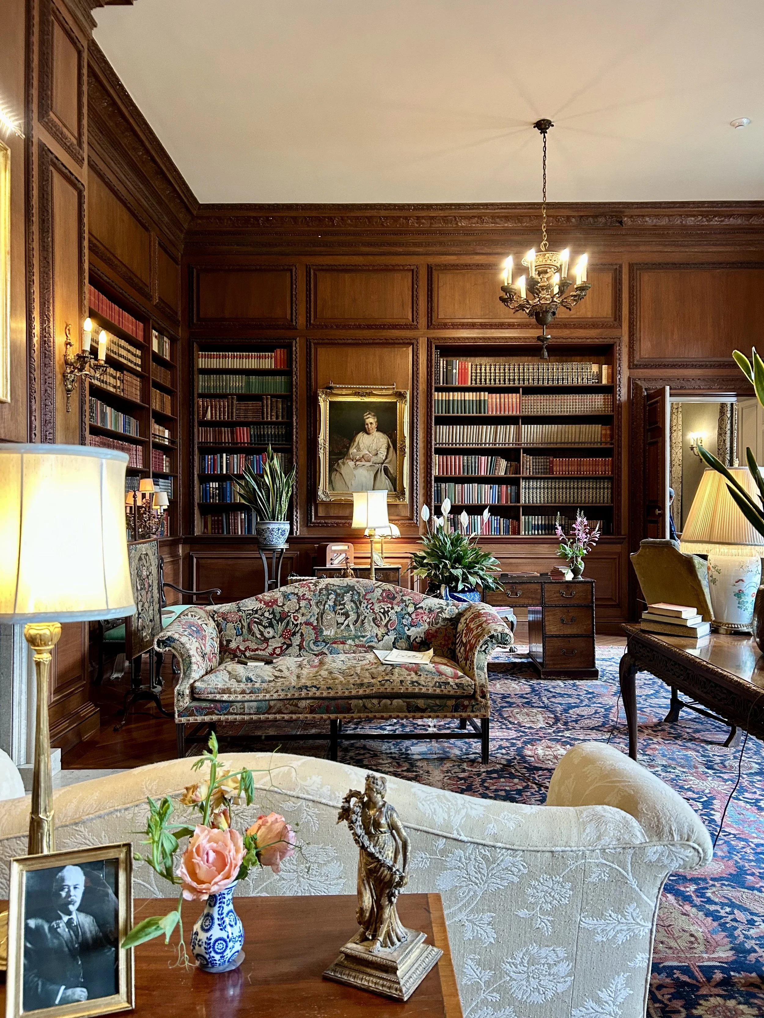 A luxurious sitting room with wooden paneled walls, filled with bookshelves, vintage furniture, and decorative items. There is a floral-patterned sofa, a small wooden table with a framed black-and-white portrait, a vase with pink flowers, and a golde