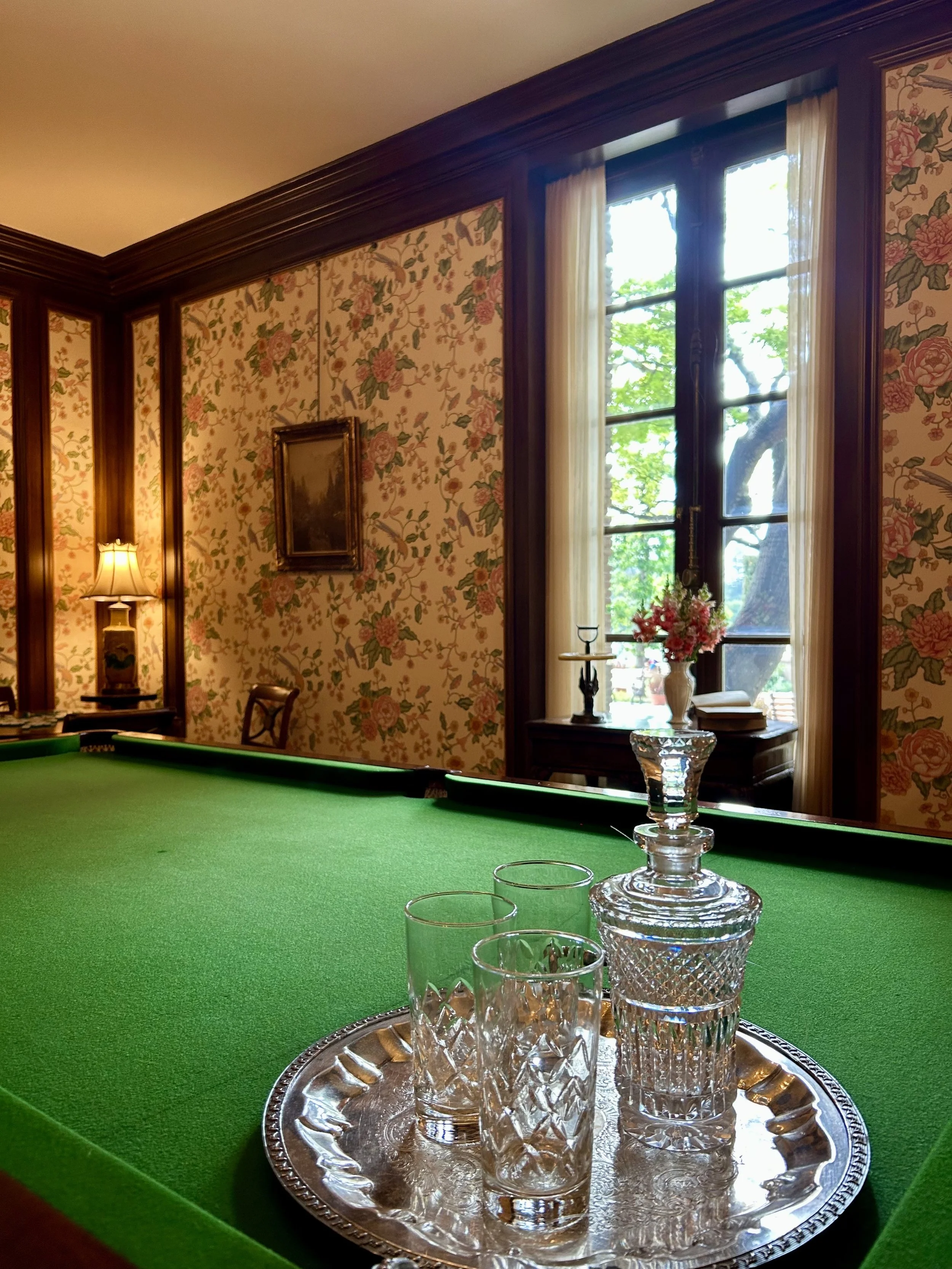 A room with floral wallpaper, a window with white curtains, a small table with pink flowers, and a pool table with a green felt surface. On the table, there is a silver tray with glassware, including a decanter and glasses. The room has dark wood tri
