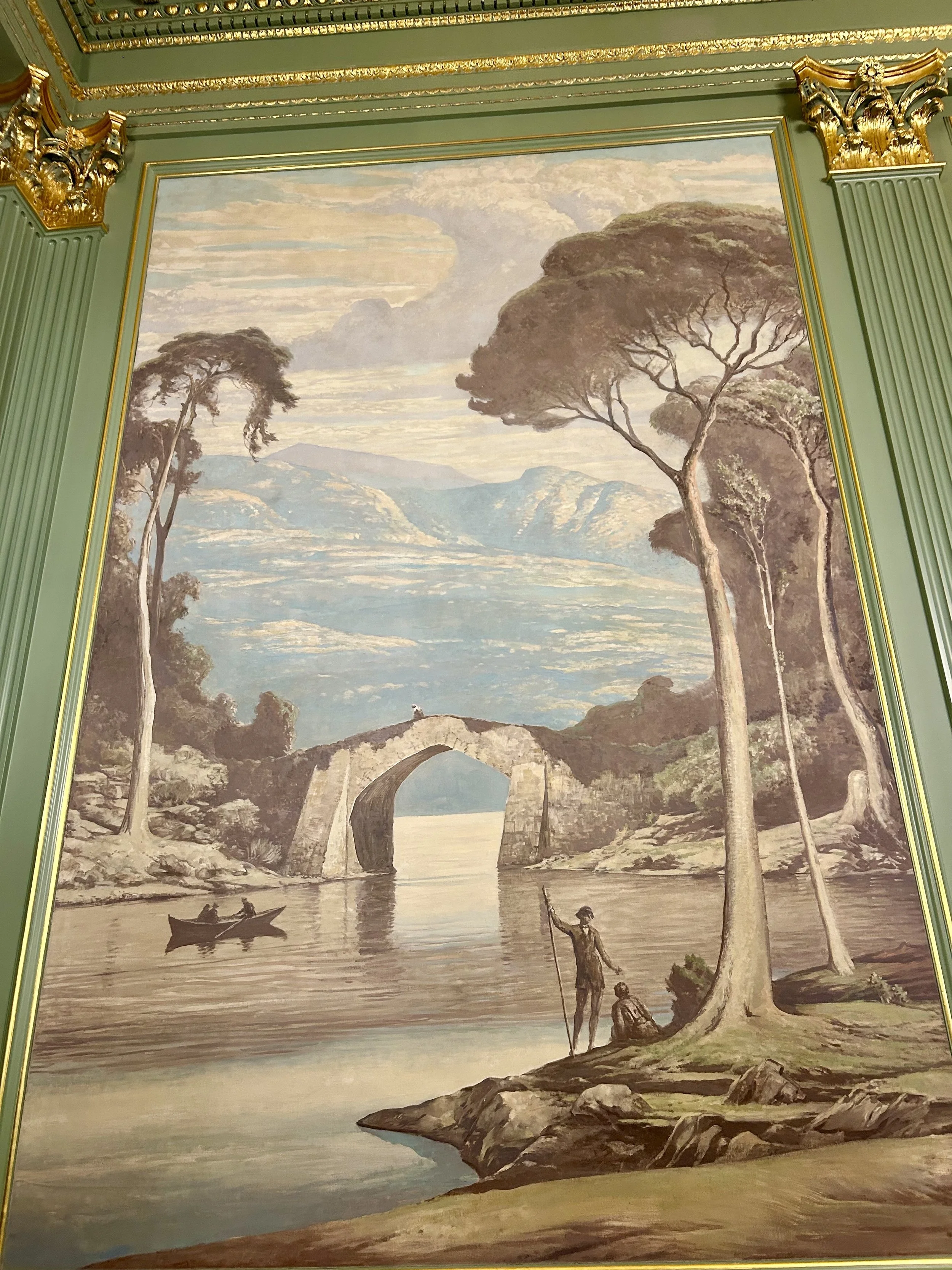 A landscape painting featuring trees, mountains, a river, and people in boats near a stone arch bridge.