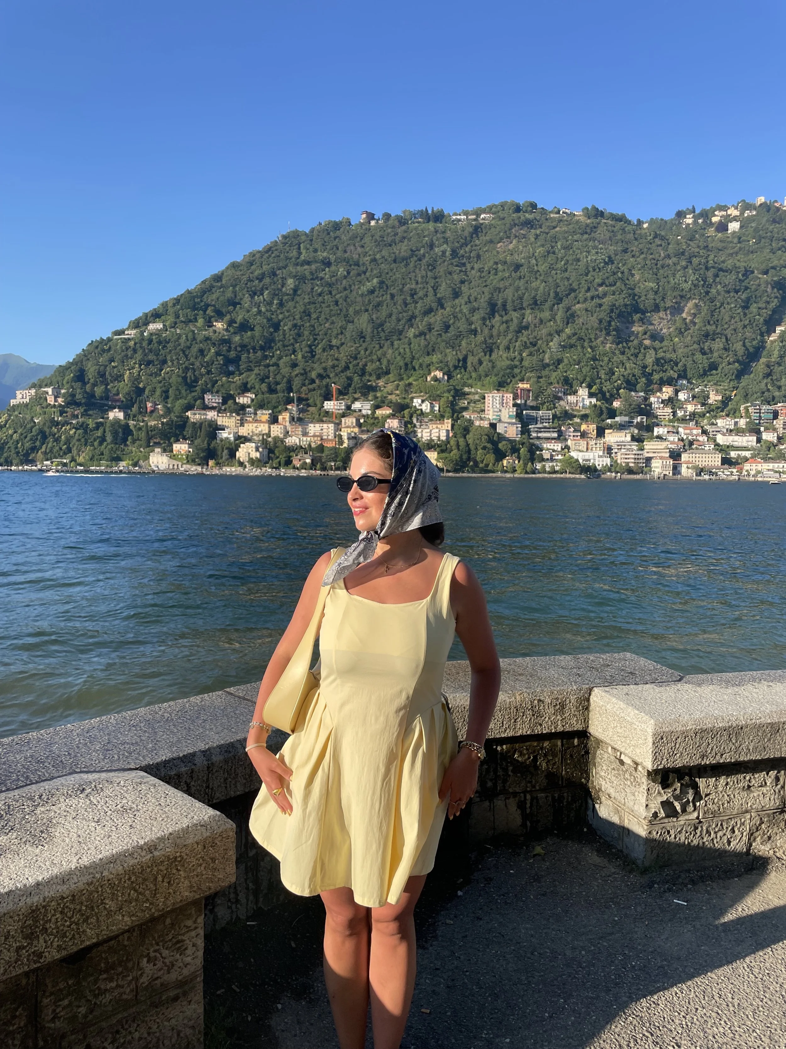 A woman with a yellow dress standing by a stone railing near a large body of water with a hill covered with trees and buildings in the background, sunglasses, and a scarf over her hair.