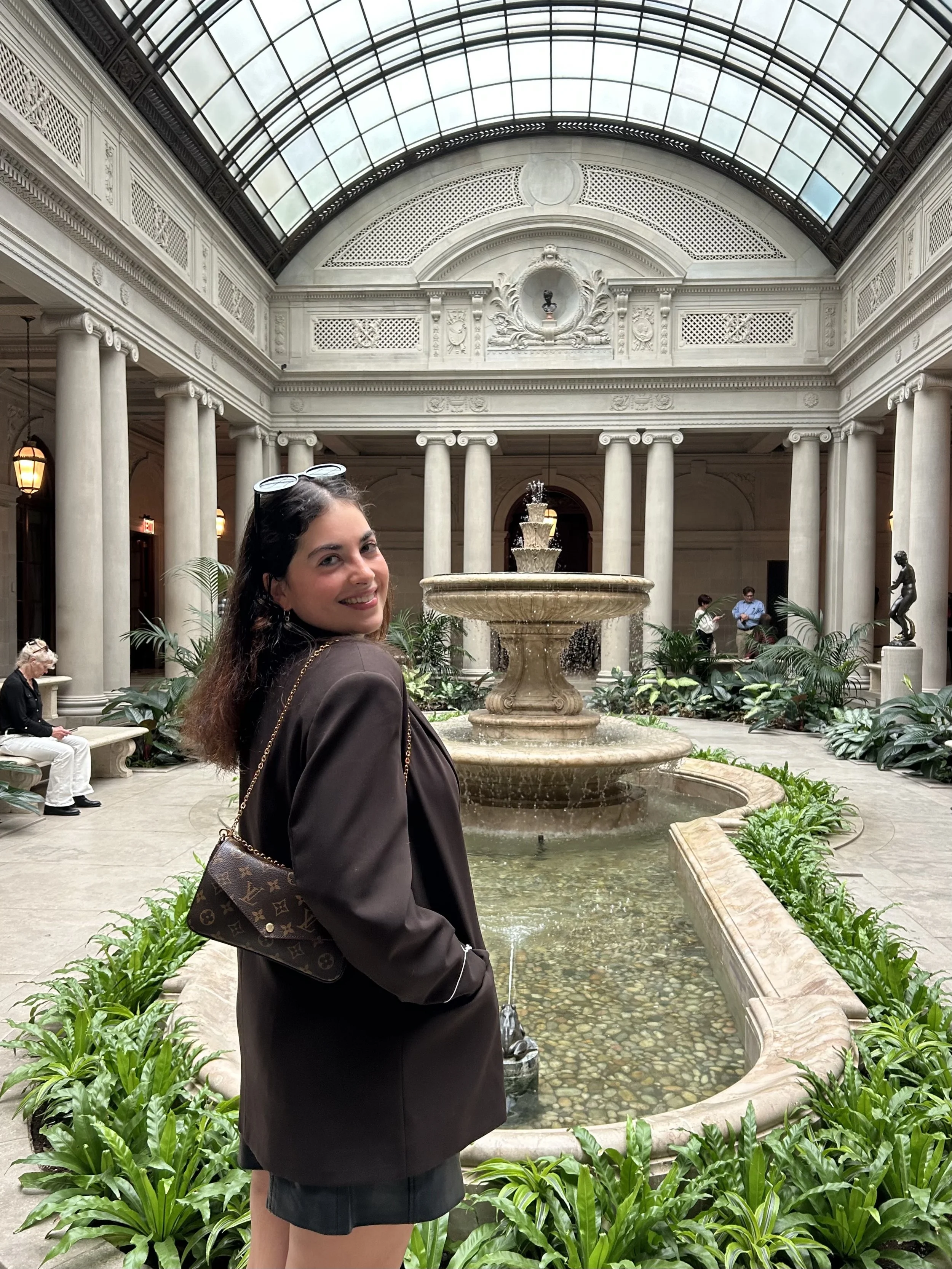 A woman with glasses on her head, smiling, standing by a fountain in a grand indoor space with marble columns and a glass ceiling.