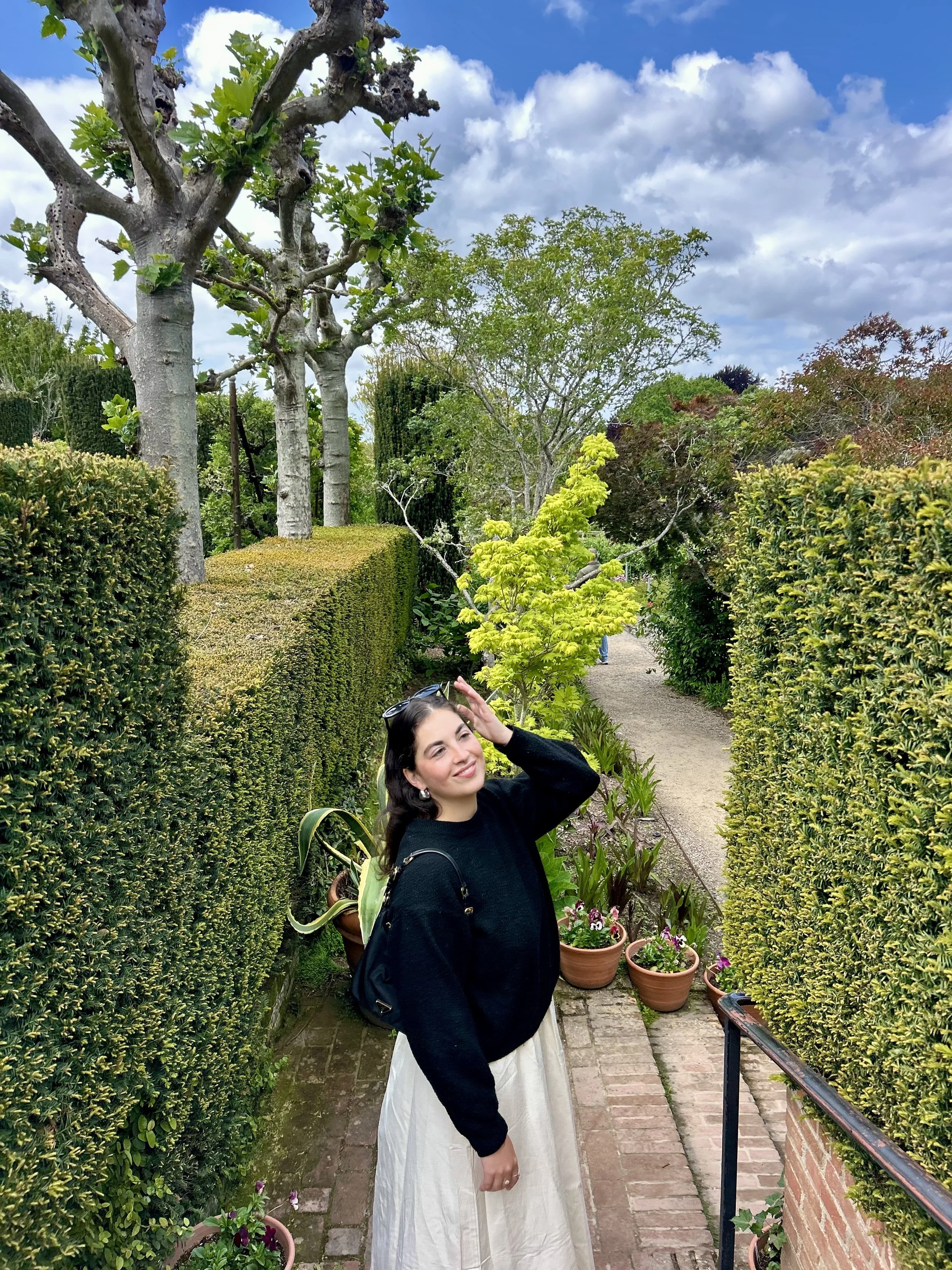 A smiling woman with long dark hair, wearing a black sweater and flowing white skirt, standing on a garden pathway surrounded by neatly trimmed green hedges and potted plants, with a partly cloudy sky overhead.